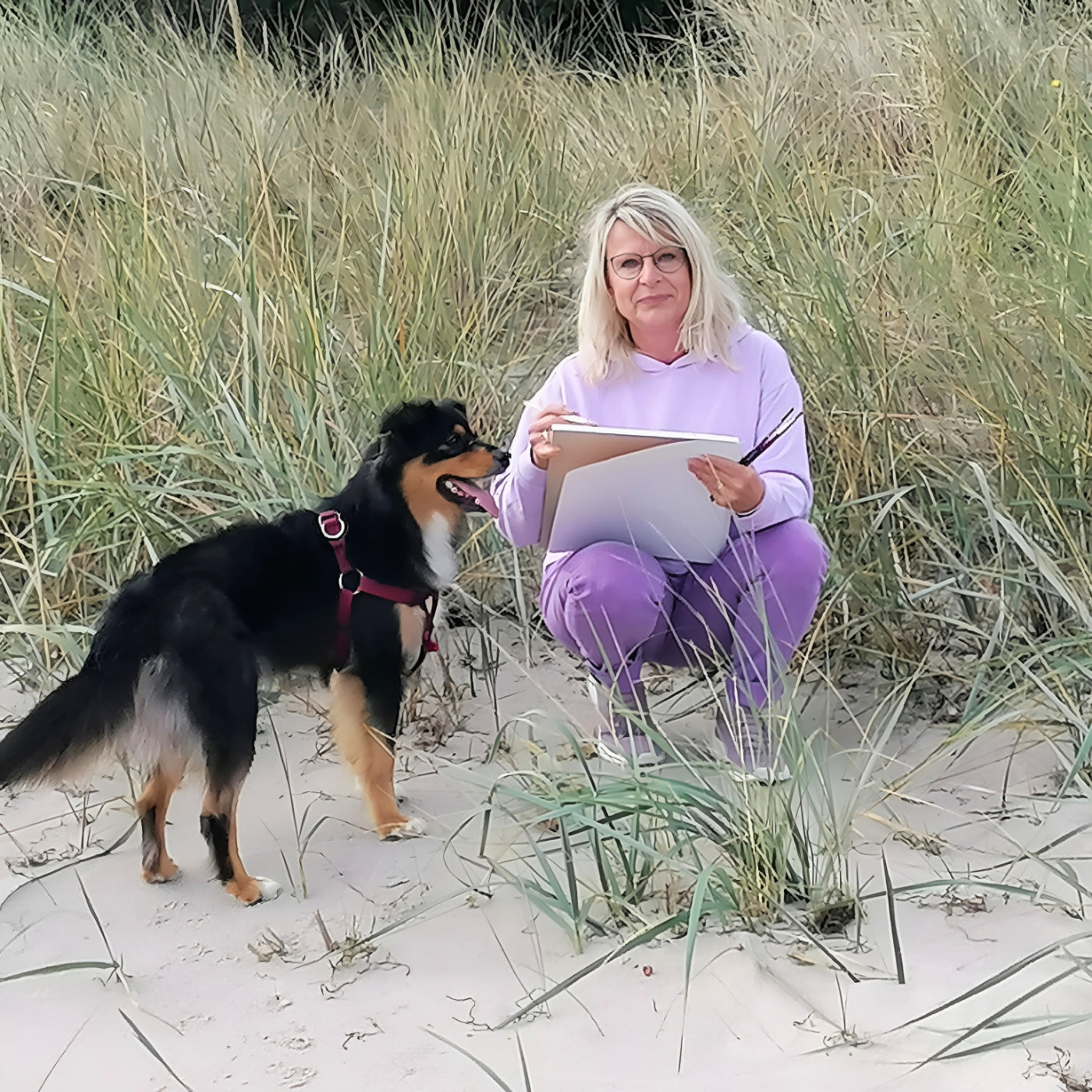 Person zeichnet im Dünengras am Strand, neben ihr steht ein Hund; ruhige Outdoorszene mit Skizzenpapier und Naturkulisse.
