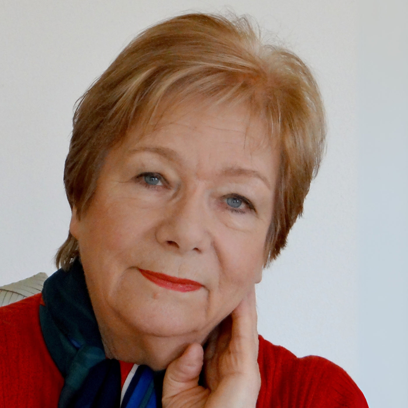 Anke Huober, Künstlerin der Online-Galerie FemArtSPACE, lächelt leicht. Die Frau mittleren Alters mit kurzen blonden Haaren trägt einen roten Pullover und einen blau-grün gemusterten Schal. Ihr Kinn ruht auf ihrer rechten Hand. 