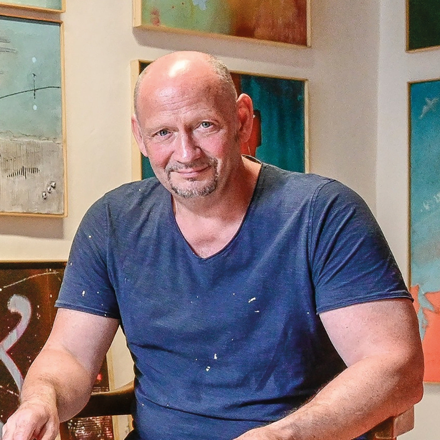 Künstler im Atelier, vor mehreren abstrakten Acrylwerken sitzend, mit Farbflecken auf Kleidung und einer entspannten Körperhaltung.