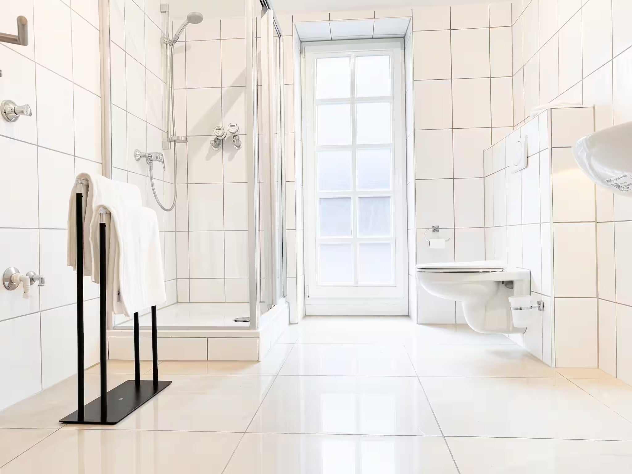 Ein modernes, hell gefliestes Badezimmer im Gutshaus am Rhein (Duisburger Landstraße 37, 40489 Düsseldorf). Das Bad der Art Apartments verfügt über eine gläserne Duschkabine, ein wandhängendes WC und ein großes Sprossenfenster, das für viel Tageslicht sorgt. Ein schwarzer, minimalistischer Handtuchhalter setzt einen eleganten Akzent. Diese hochwertig ausgestatteten Zimmer können von Reisenden sowie für exklusive Ausstellungen gebucht werden.