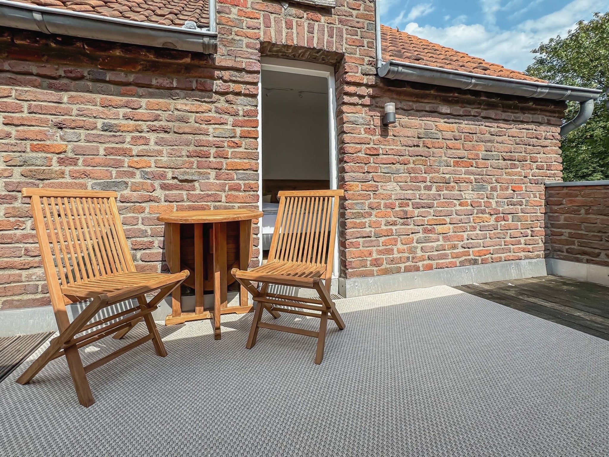 Eine einladende Dachterrasse mit rustikaler Backsteinwand im Gutshaus am Rhein (Duisburger Landstraße 37, 40489 Düsseldorf). Zwei Klappstühle und ein kleiner Tisch aus hellem Holz stehen auf einem hellgrauen Außenteppich vor dem Eingang zu einem der modernen Zimmer. Diese Art Apartments bieten eine einzigartige Atmosphäre für Reisende sowie Raum für Ausstellungen und können direkt gebucht werden.