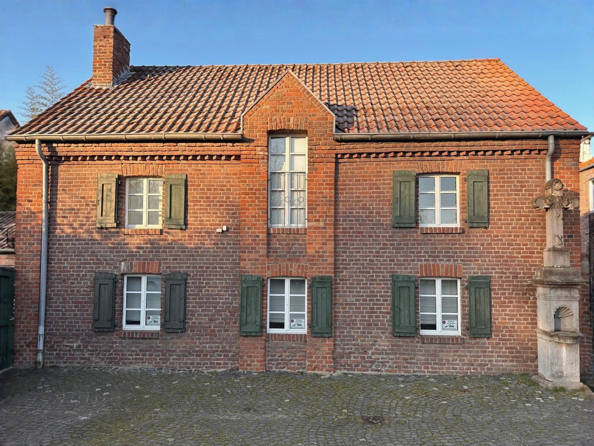 Die historische Backsteinfassade des Gutshauses am Rhein (Duisburger Landstraße 37, 40489 Düsseldorf) bei sonnigem Wetter. Das Gebäude der Art Apartments beeindruckt durch seine klassische Architektur mit grünen Fensterläden, einem roten Ziegeldach und einem markanten Steinkreuz im Vordergrund. Die stilvollen Apartments und Zimmer stehen für Reisende zur Buchung bereit und bieten zudem einen exklusiven Rahmen für Ausstellungen.