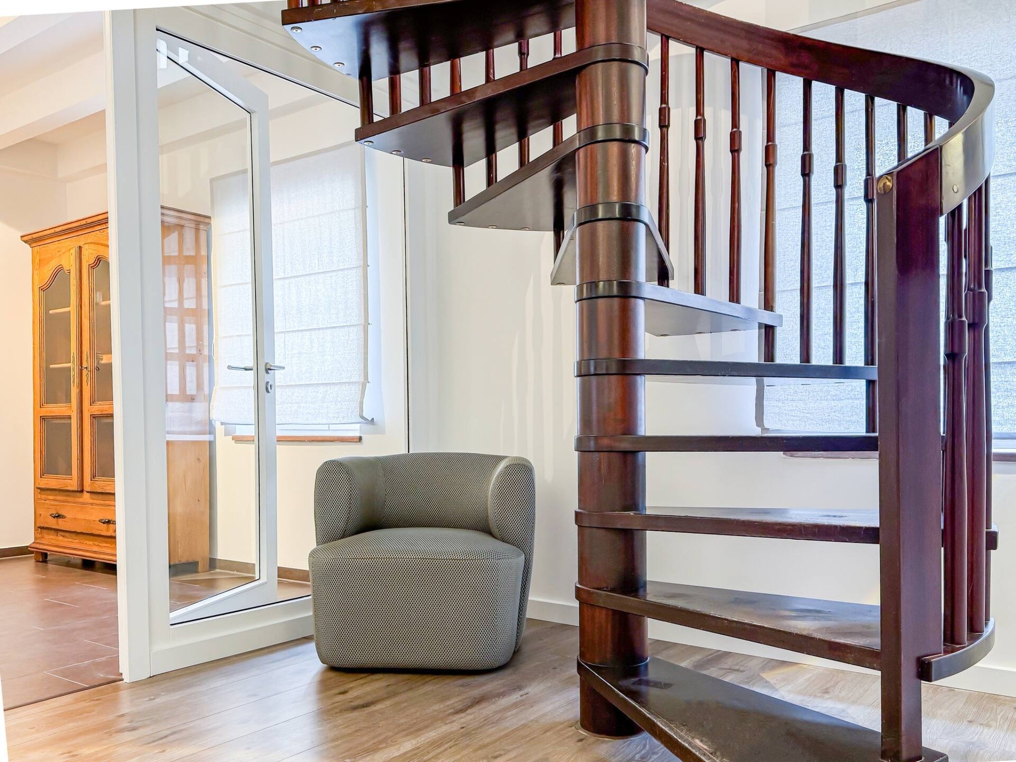 Detailansicht einer eleganten, dunklen Holzwendeltreppe im Gutshaus am Rhein (Duisburger Landstraße 37, 40489 Düsseldorf). Ein moderner, grauer Sessel steht einladend neben der Treppe auf dem hellen Holzboden, während im Hintergrund ein antiker Holzschrank durch eine Glastür sichtbar ist. Die Art Apartments bieten eine harmonische Mischung aus Design und Historie für Reisende und können zudem für Ausstellungen von Künstlern der Online-Galerie ARTSPACER gebucht werden.