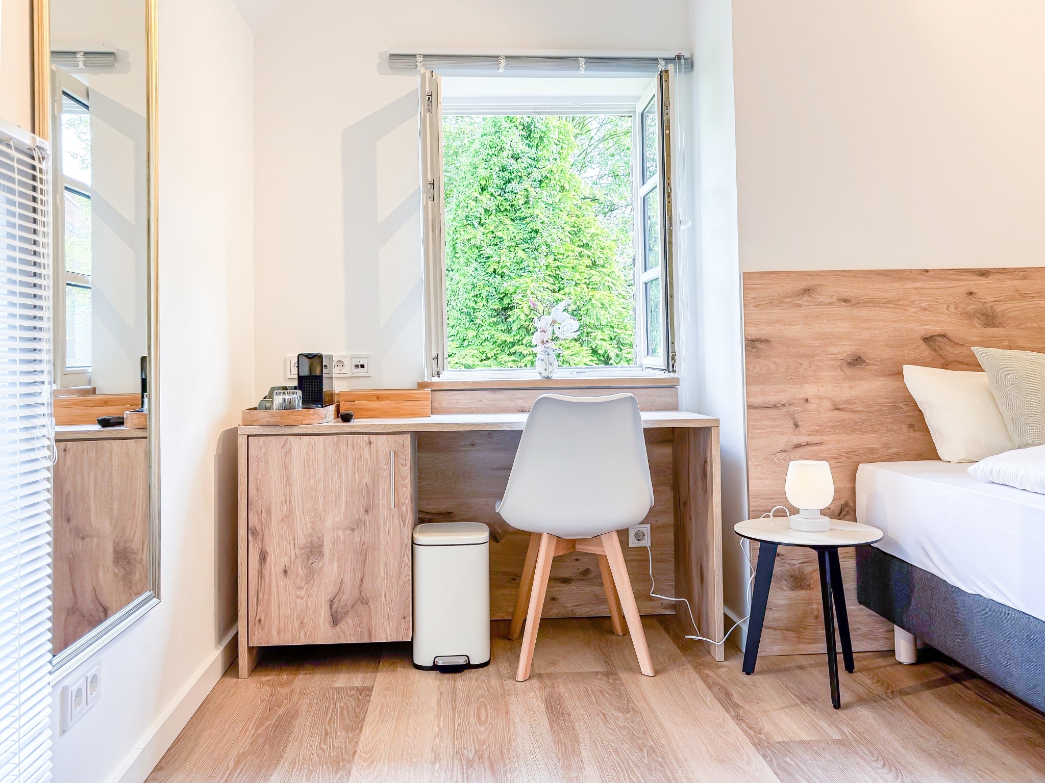 Ein lichtdurchfluteter Arbeitsplatz in einem der Zimmer im Gutshaus am Rhein (Duisburger Landstraße 37, 40489 Düsseldorf). Der Schreibtisch aus hellem Holz ist direkt unter einem Fenster mit Blick ins Grüne platziert. Daneben befindet sich ein gemütliches Boxspringbett mit Holz-Kopfteil und ein moderner Nachttisch mit Lampe. Diese stilvollen Art Apartments bieten eine ideale Umgebung für Reisende sowie für kreative Ausstellungen und können gebucht werden.