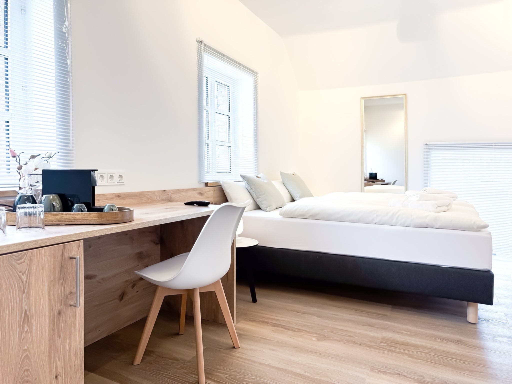 Ein kombinierten Schlaf- und Arbeitsbereich in einem modernen Zimmer im Gutshaus am Rhein (Duisburger Landstraße 37, 40489 Düsseldorf). Im Vordergrund steht ein Schreibtisch aus hellem Holz mit einem weißen Designerstuhl und einer Kaffeestation, dahinter ein bequemes Boxspringbett mit frischer weißer Wäsche. Die Art Apartments bieten eine helle Wohlfühlatmosphäre für Reisende und können zudem für Ausstellungen  für Künstler der Online-Galerie ARTSPACER gebucht werden.