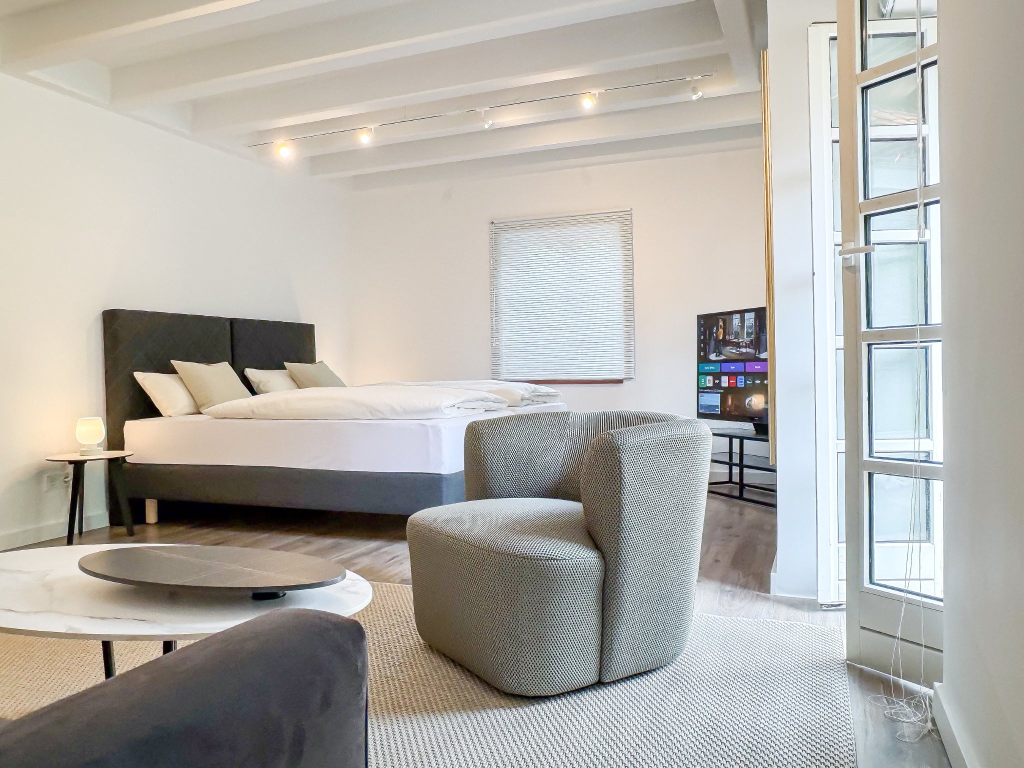 Der modern gestaltete Wohn- und Schlafbereich im Gutshaus am Rhein (Duisburger Landstraße 37, 40489 Düsseldorf). Im Vordergrund steht ein strukturierter grauer Sessel neben einem Marmor-Couchtisch, im Hintergrund ist ein großes Doppelbett und ein Smart-TV unter einer weißen Balkendecke zu sehen. Die Art Apartments bieten ein exklusives Ambiente für Reisende und können für Ausstellungen von Künstlern der Online-Galerie ARTSPACER gemietet werden.