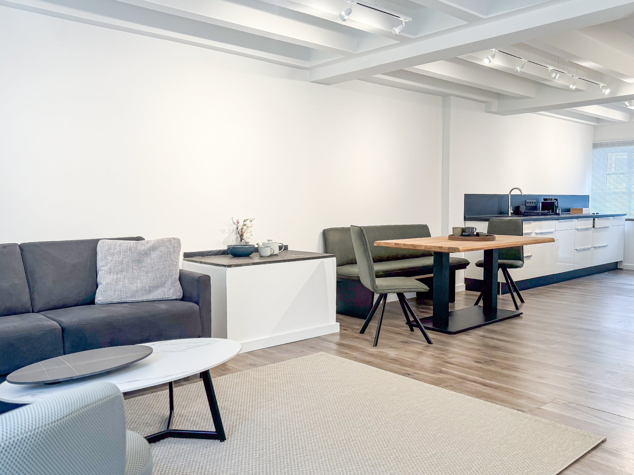Der großzügige, offen gestaltete Wohn- und Essbereich eines Art Apartments im Gutshaus am Rhein (Duisburger Landstraße 37, 40489 Düsseldorf). Zu sehen ist ein gemütliches graues Sofa mit Couchtisch, ein massiver Esstisch mit olivgrüner Sitzbank und die angrenzende, moderne weiße Küchenzeile. Die helle Raumgestaltung mit weißer Balkendecke und moderner Spot-Beleuchtung bietet eine ideale Atmosphäre für Reisende sowie für exklusive Ausstellungen von Künstlern der Online-Galerie ARTSPACER.