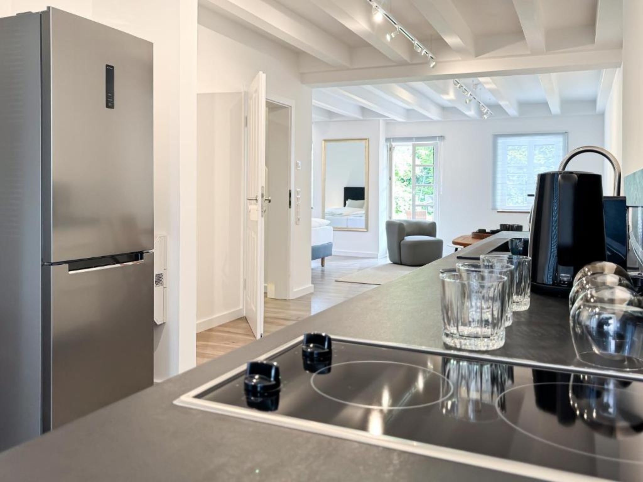 Blick in die moderne, offene Küche und den Wohnbereich eines Art Apartments im Gutshaus am Rhein (Duisburger Landstraße 37, 40489 Düsseldorf). Die Ausstattung umfasst ein Cerankochfeld, Gläser und einen Edelstahl-Kühlschrank im Vordergrund, während im Hintergrund der helle Wohnraum mit weißer Balkendecke, Sessel und Bett zu sehen ist. Diese stilvollen Unterkünfte können von Reisenden sowie für Ausstellungen und Kulturevents gebucht werden.