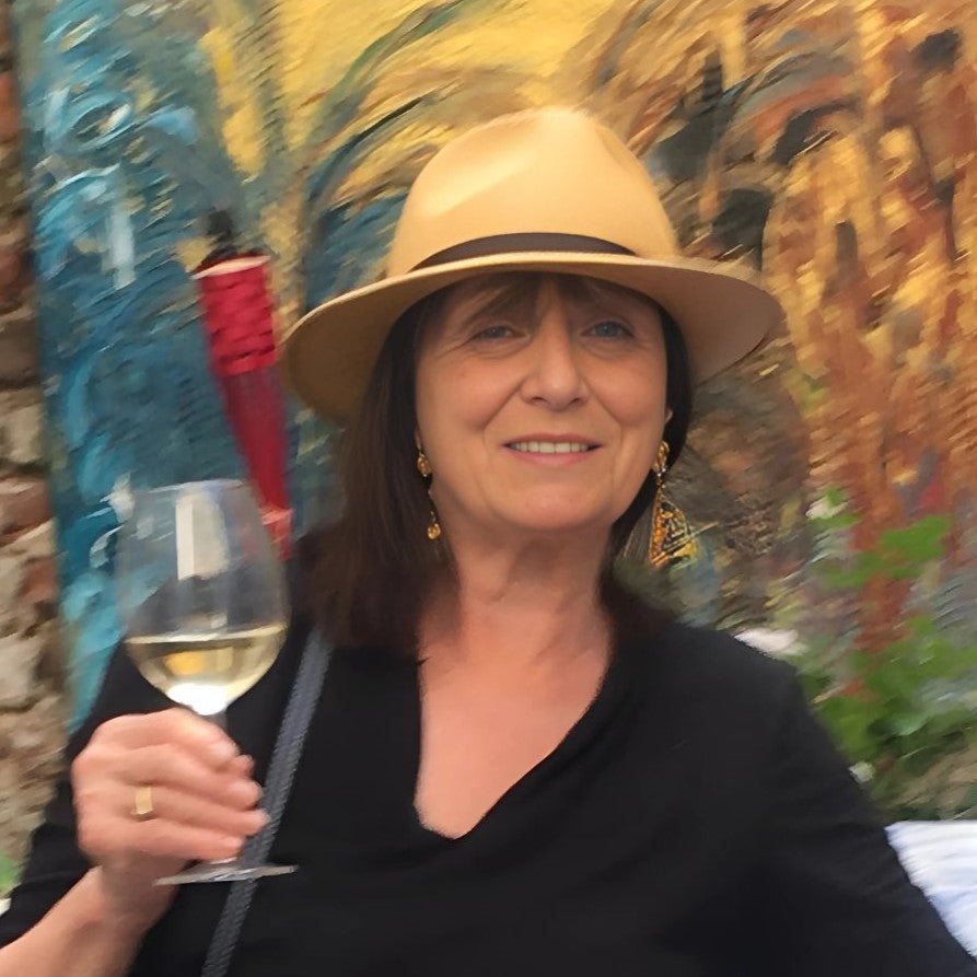 Porträt der Künstlerin Federica aus Düsseldorf (FEMARTSPACE): Eine freundlich lächelnde Frau mit braunen Haaren und hellen Augen trägt einen hellen Hut mit dunklem Band und auffällige, goldfarbene Ohrringe. Sie hält in ihrer rechten Hand ein Glas mit einem hellen Getränk. Der Hintergrund ist unscharf und zeigt eine Wand mit bunten, abstrakten Malereien in Blau-, Gelb- und Brauntönen.