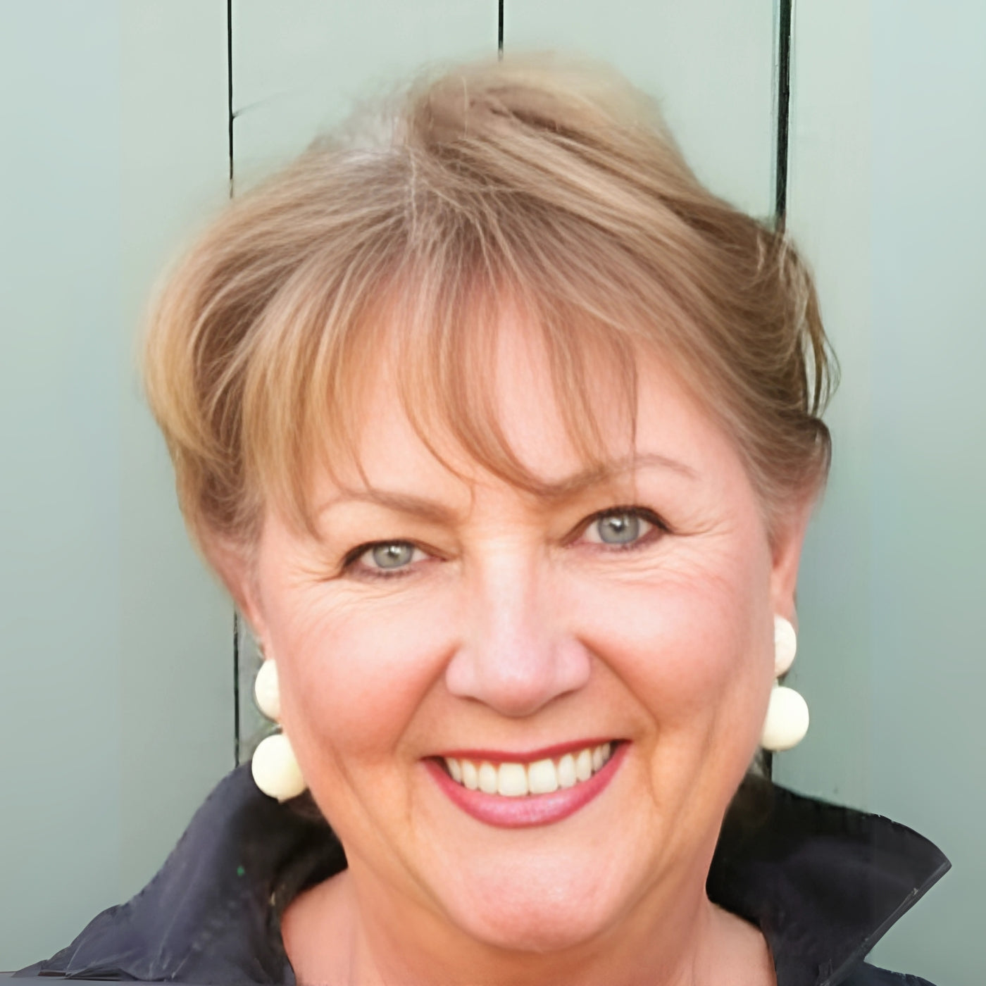 Porträt von Claudia Birkheuer (FEMARTSPACE): Ein Nahaufnahme-Porträt der Künstlerin. Sie hat kurze, gewellte, blonde Haare mit grauen Strähnen und strahlend blaue Augen. Sie lächelt freundlich und zeigt ihre Zähne. Sie trägt große, runde, cremefarbene Ohrringe und eine dunkle Jacke mit hochgeschlagenem Kragen. Der Hintergrund ist eine hellgrüne, vertikal geriffelte Oberfläche.