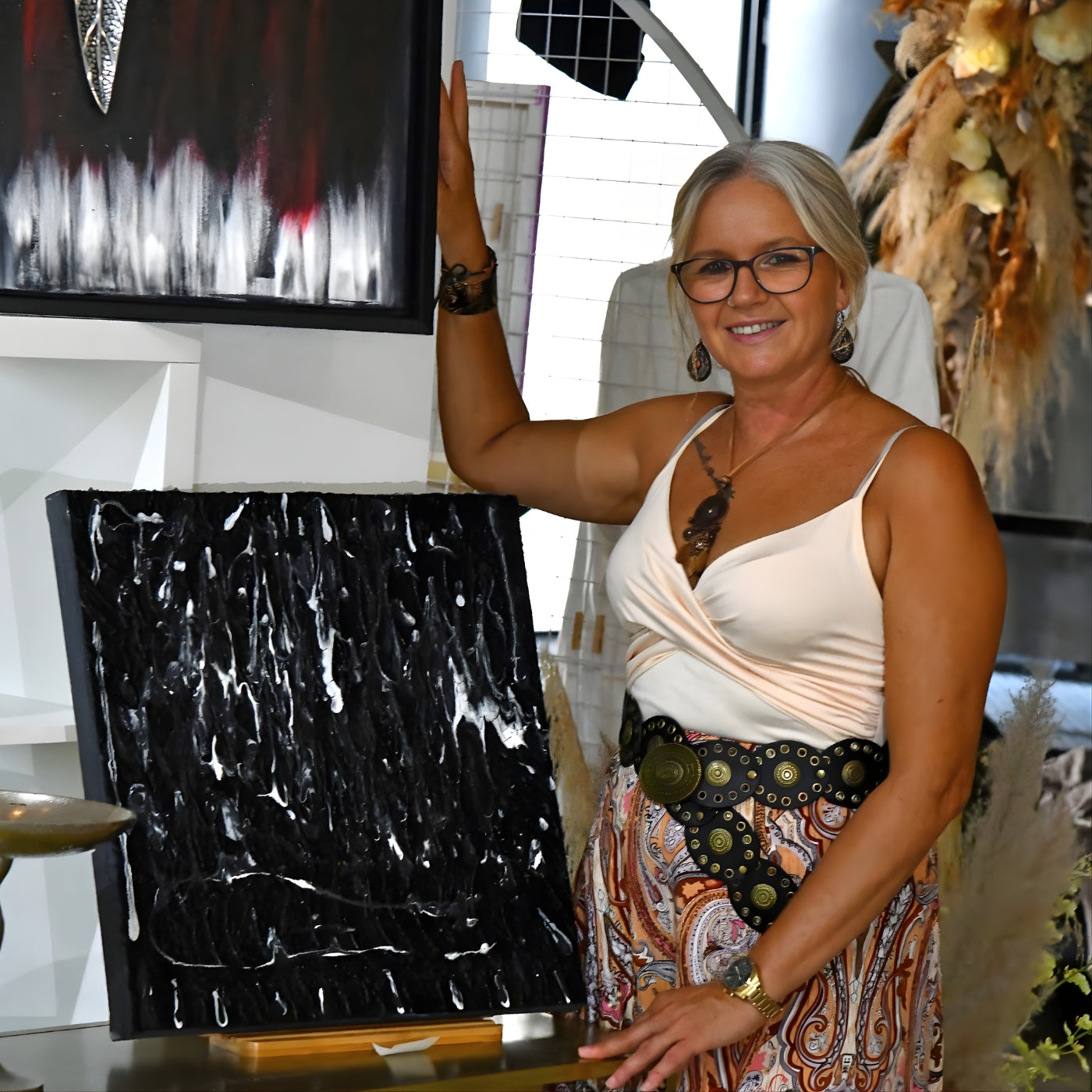 Porträt der Künstlerin Sandra Conrad, die lächelnd neben zwei ihrer abstrakten Kunstwerke steht. Sie hat graue Haare, trägt eine Brille und ist in einem hellen Oberteil und einem gemusterten Rock zu sehen. Rechts neben ihr steht ein quadratisches, strukturiertes Werk in Schwarz und Weiß, mit dicken, tropfenartigen Linien. Ein weiteres Gemälde im Hintergrund zeigt rote und weiße Akzente auf schwarzem Grund.