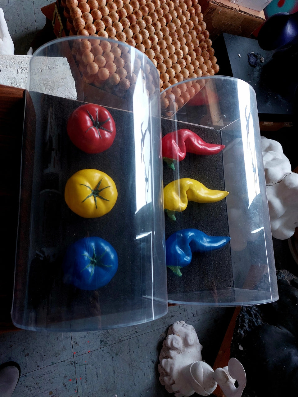 Skulptur von Amos Plaut: Zwei zylinderförmige, transparente Kunststoffbehälter stehen nebeneinander auf einem grauen, unebenen Untergrund. In jedem Behälter befinden sich drei stilisierte, organisch geformte Objekte in den Farben Rot, Gelb und Blau. Die Objekte im linken Behälter sind kompakter und ähneln stilisierten Tomaten. Die Objekte im rechten Behälter sind länglicher und gewundener, mit einer tropfenartigen Form. Im Hintergrund sind unscharf weitere Ateliergegenstände zu erkennen.