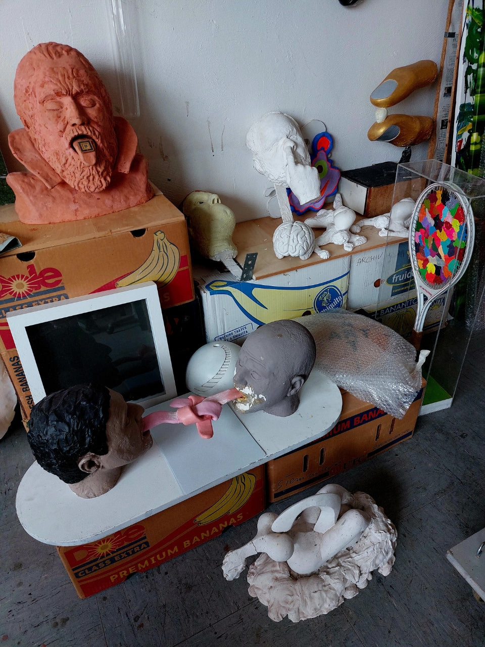 Atelier von Amos Plaut: Ein unaufgeräumter Arbeitsbereich voller unfertiger und fertiger Kunstwerke. Auf Kartons und einer weißen Platte liegen verschiedene Skulpturen aus unterschiedlichen Materialien wie Ton und Gips, darunter mehrere Kopfstudien mit expressiven Gesichtszügen. Ein rotbrauner, fragmentierter Kopf mit aufgerissenem Mund liegt auf einem Bananenkarton. Ein weiterer, grauer Kopf ist auf einer weißen Unterlage platziert. Im Hintergrund lehnen weitere Objekte an einer weißen Wand, 