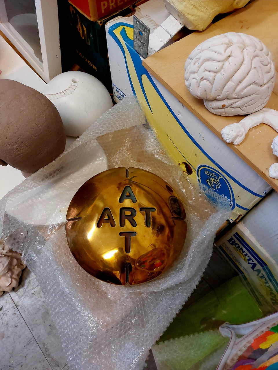 Installation 'Pille für die Kunst' von Amos Plaut: Eine glänzende, goldfarbene, runde Dose oder Schale liegt auf einer zerknitterten, durchsichtigen Folie. In die Oberfläche der Dose sind die Buchstaben 'ART' in einer vertikalen Anordnung eingeprägt, wobei das 'A' oben und das 'T' unten steht. Um die goldene Dose herum liegen verschiedene andere Objekte, darunter ein weißes, geformtes Objekt, das an ein Gehirn erinnert, Fragmente von Kartons mit Bananenaufdruck.