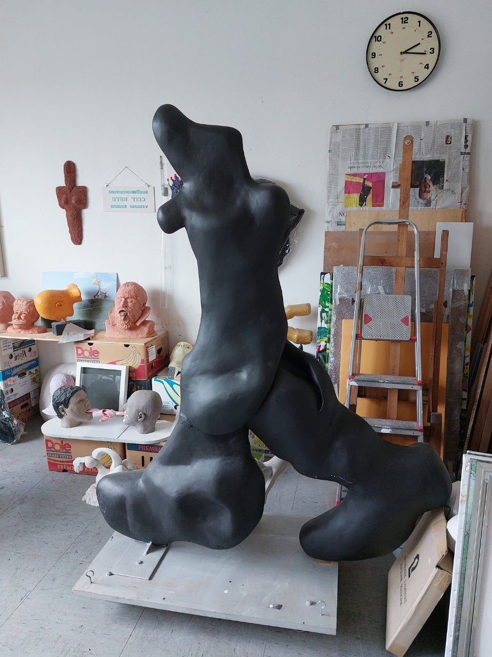 Skulptur von Amos Plaut: Eine große, organisch geformte Skulptur aus dunklem Material, möglicherweise Ton oder Gips, dominiert den Raum eines Ateliers. Die Skulptur erinnert an einen stilisierten, liegenden oder sich windenden Körper mit abgerundeten Formen und Ausstülpungen. Sie steht auf einer hellen, rechteckigen Basis. Im Hintergrund ist ein weiß gestrichener Raum mit verschiedenen weiteren Kunstwerken, Skulpturen auf Regalen, einer Leiter, Zeitungen an der Wand & einer runden Uhr zu sehen