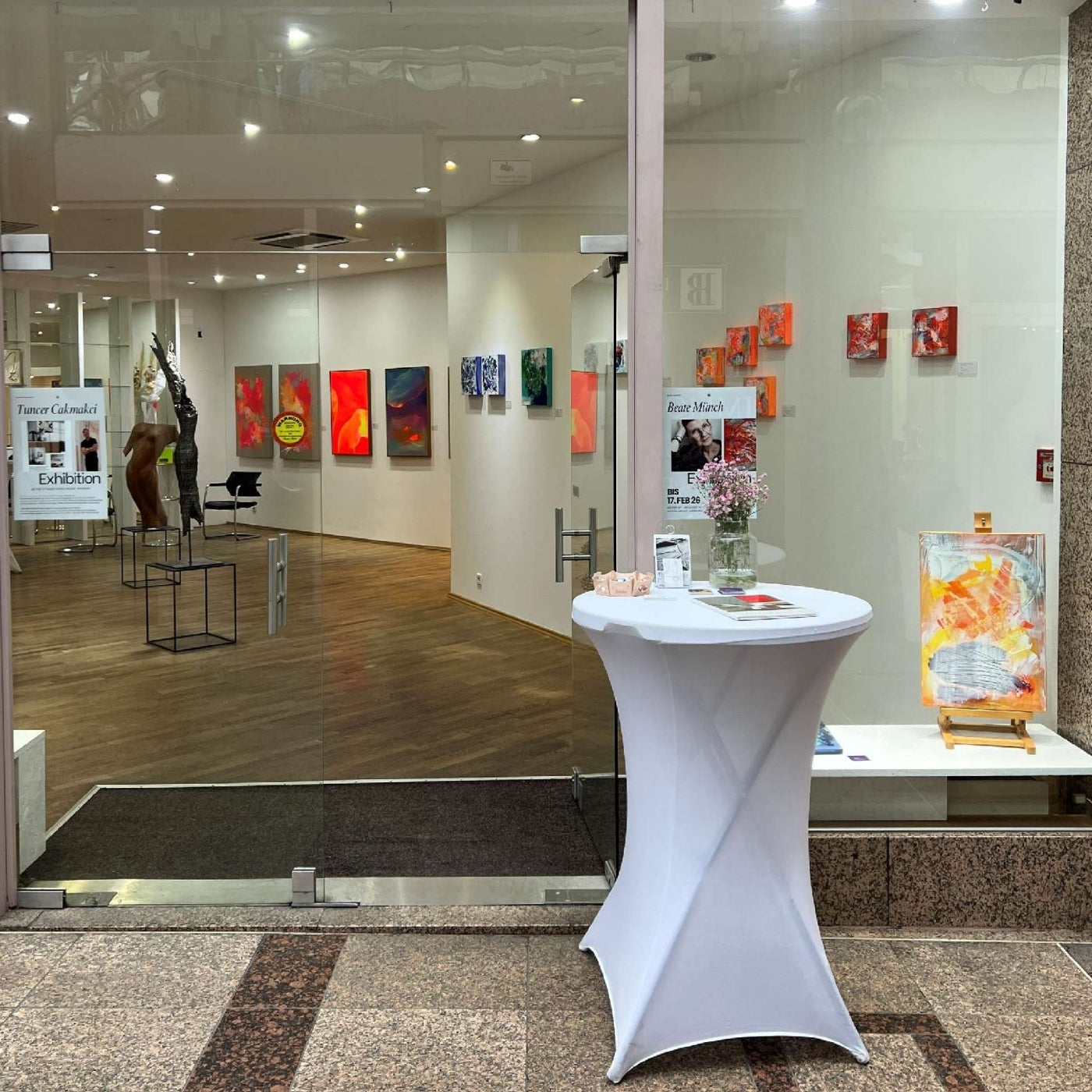 Blick durch die Glasfront in die ART Pop-Up Galerie in der Wilhelm-Arcade (Wilhelmstraße 38, Wiesbaden). Die hellen Galerieräume zeigen moderne Gemälde in kräftigen Rot- und Blautönen sowie abstrakte Skulpturen auf schwarzen Sockeln. Im Vordergrund steht ein weiß eingekleideter Stehtisch mit Infomaterial. Künstler der Online-Galerie ARTSPACER können sich hier für physische Ausstellungen bewerben, um ihre Werke in bester Lage zu präsentieren.