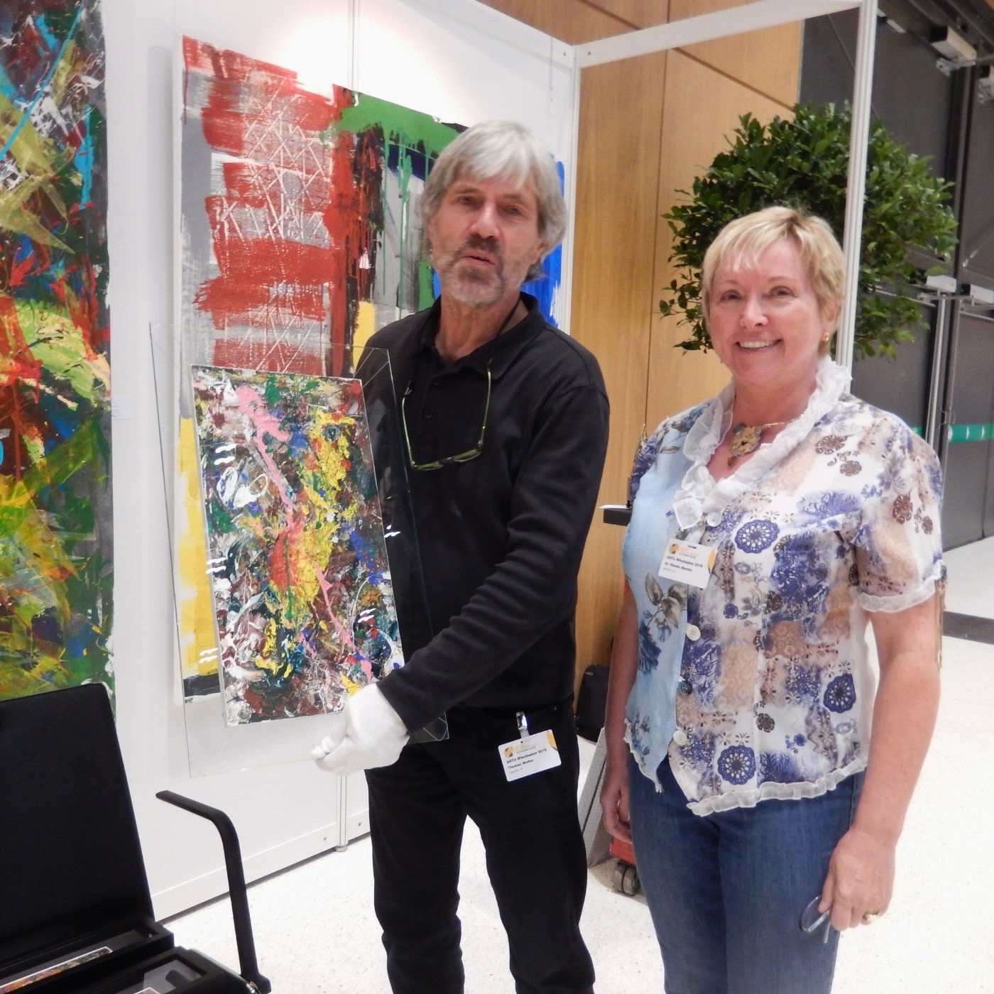 Die Künstlerin Blanka Mandel auf der Messe ARTE Wiesbaden. Neben ihr hält ein Mann mit weißen Handschuhen eines ihrer farbenfrohen, abstrakten Werke auf Glas. Im Hintergrund ist ein weiteres großformatiges Gemälde der Künstlerin zu sehen.
