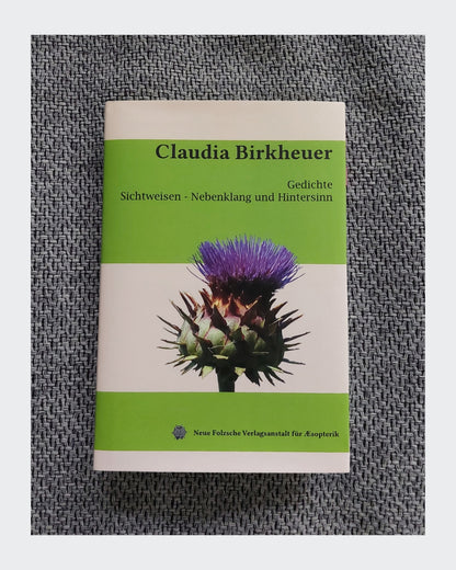 
Ein Buchcover mit dem Titel „Claudia Birkheuer Gedichte: Sichtweisen - Nebenklang und Hintersinn.“ Das Cover ist in drei horizontale Abschnitte unterteilt: oben grün, eine hellgraue oder cremefarbene Mitte und ein grüner unterer Bereich. In der Mitte befindet sich eine Fotografie einer lila Distelblüte mit einem stacheligen, grünen Kelch.
