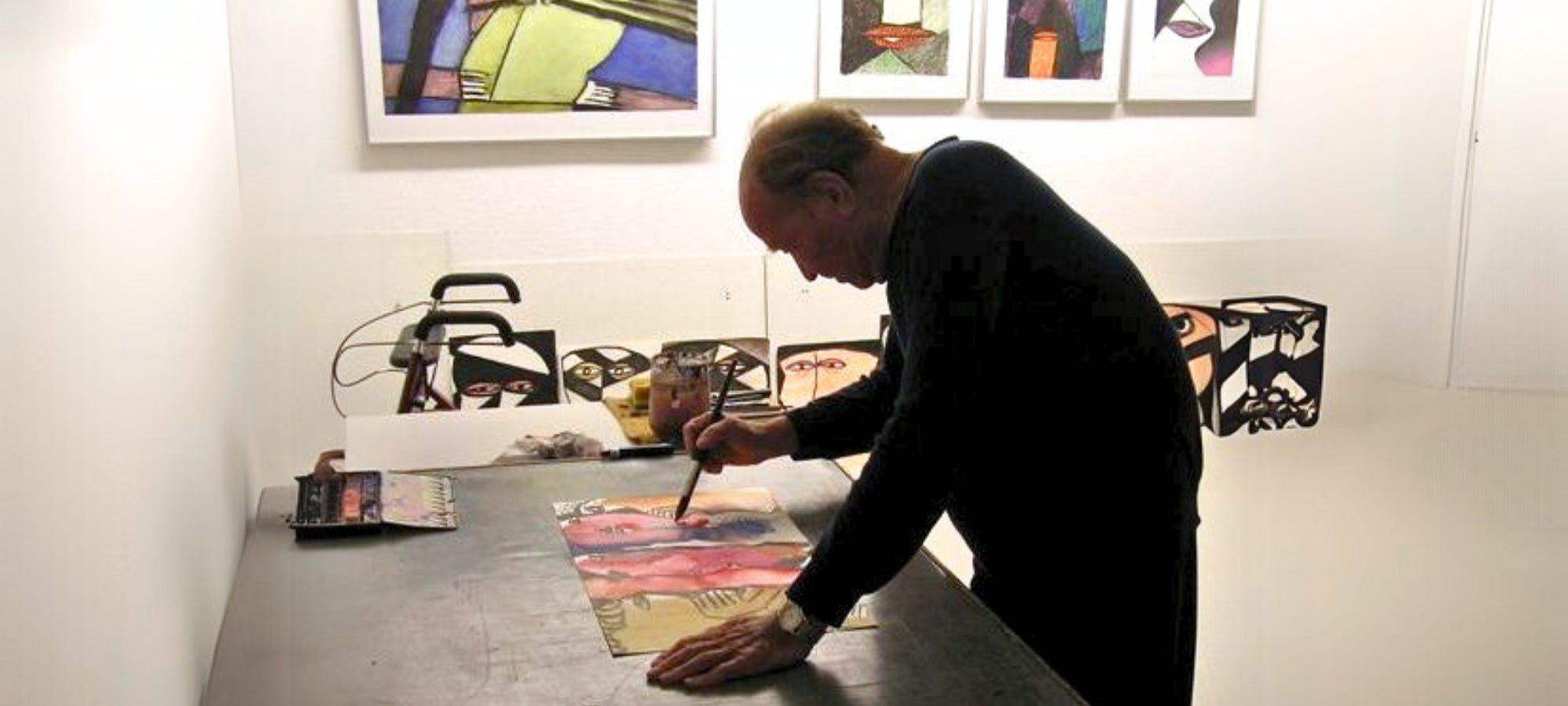 Eine authentische Aufnahme des Künstlers Franz Vornberger bei der Arbeit in seinem Atelier. Man sieht ihn in der Seitenansicht, wie er konzentriert über einen großen Arbeitstisch gebeugt mit einem Pinsel an einem farbenfrohen Werk auf Papier arbeitet. Im hellen Hintergrund hängen mehrere seiner gerahmten, expressionistischen Porträts an der Wand, während weitere fertige Arbeiten auf dem Boden und den Tischen verteilt sind. Die Szene vermittelt eine ruhige, schaffensorientierte Atmosphäre.