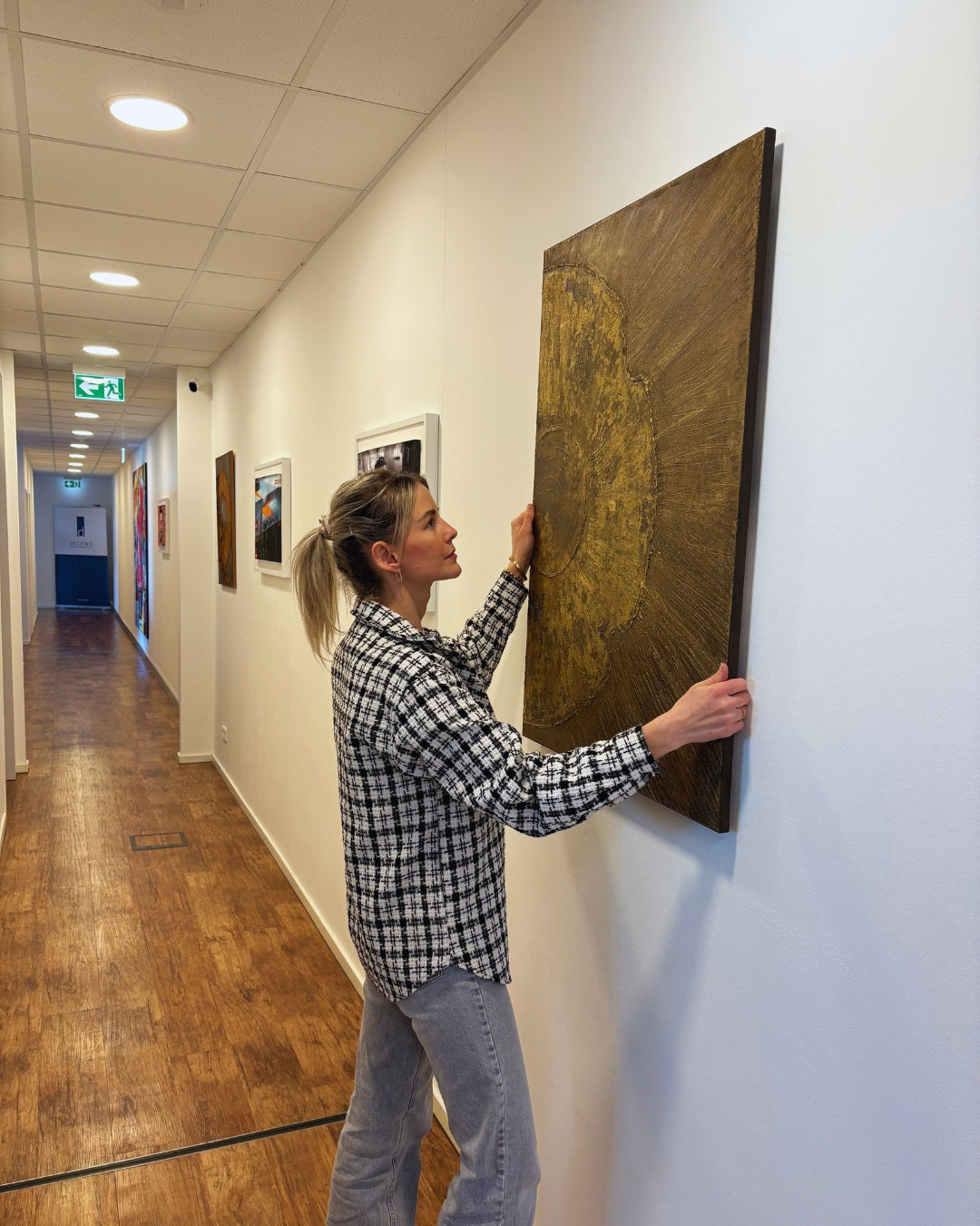Person hängt ein großformatiges abstraktes Gemälde in Gold- und Brauntönen an eine weiße Galeriewand in einem langen Flur mit Holzfußboden und Deckenbeleuchtung.