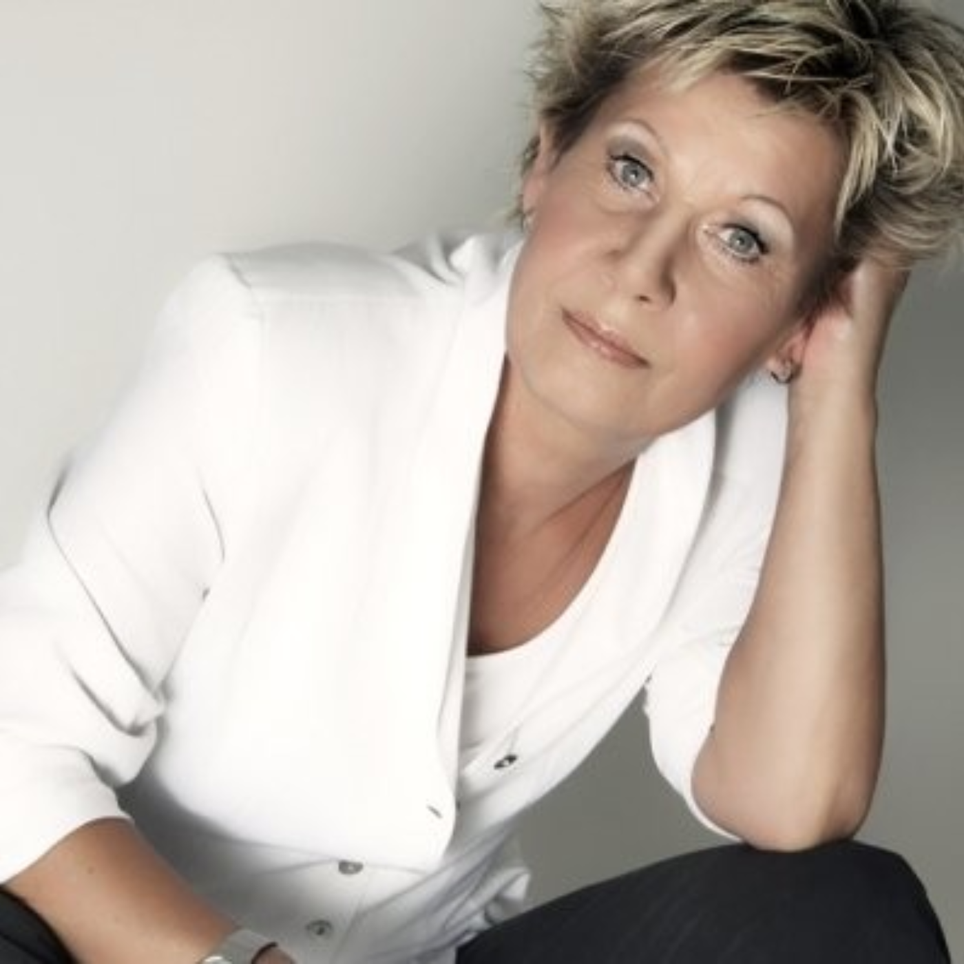 Ein Porträtfoto der Künstlerin Gabriele Springer vor einem hellen Hintergrund. Sie blickt mit einem ruhigen, nachdenklichen Ausdruck in die Kamera und stützt ihren Kopf entspannt auf ihre linke Hand. Sie trägt ihr blondes Haar kurz und leicht gestylt. Bekleidet ist sie mit einem eleganten weißen Blazer über einem passenden weißen Oberteil. Die Lichtführung ist weich und unterstreicht die professionelle, zeitlose Ausstrahlung des Porträts.