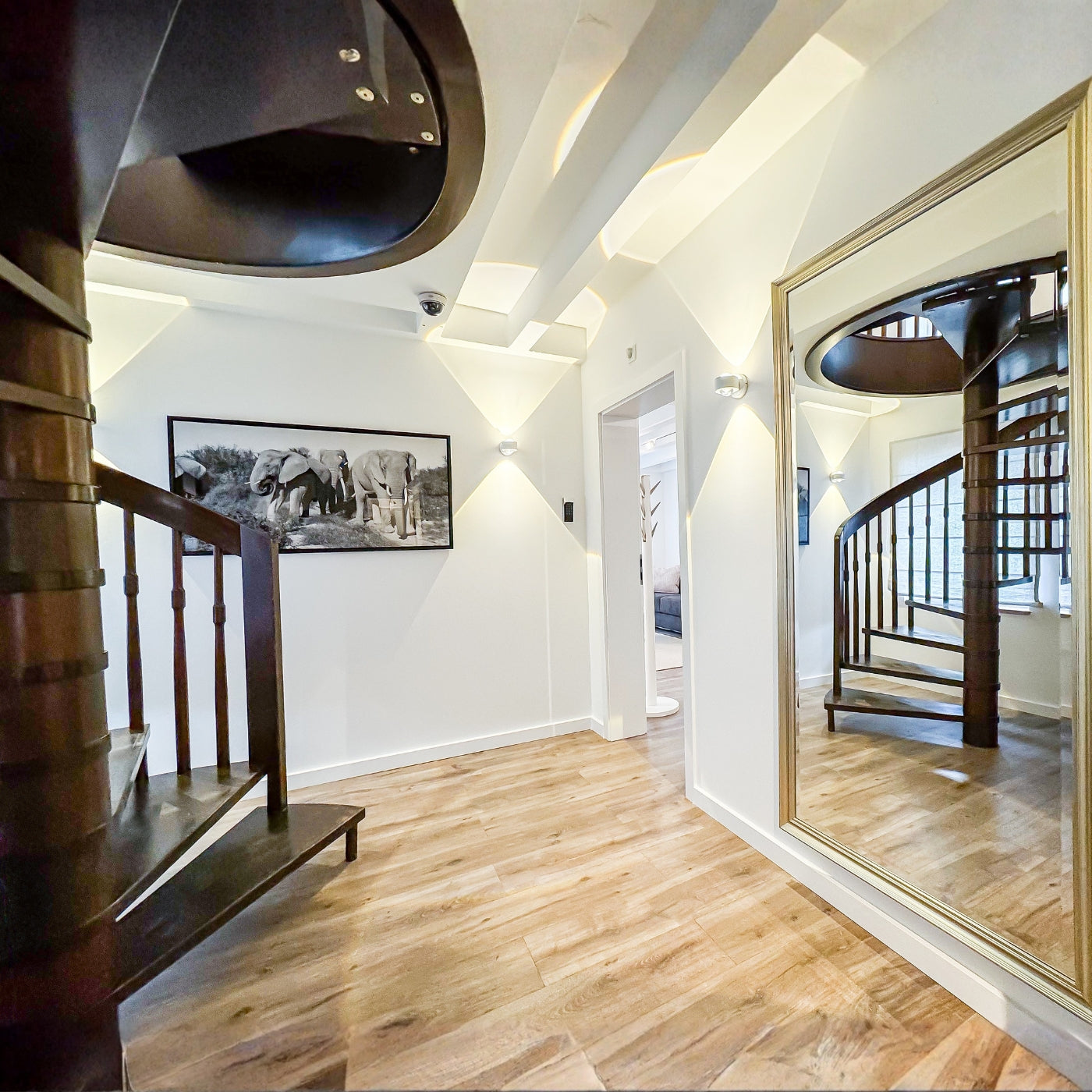 Ein heller, moderner Flur im Gutshaus am Rhein (Duisburger Landstraße 37, 40489 Düsseldorf) mit einer markanten Wendeltreppe aus dunklem Holz. Die weißen Wände werden durch Up-and-Down-Leuchten mit sanduhrförmigen Lichtkegeln in Szene gesetzt. An der Wand hängt eine Schwarz-Weiß-Fotografie von Elefanten, während ein großer Wandspiegel mit goldenem Rahmen den Raum und die Treppe reflektiert. Der Boden ist mit warmem Parkett in Holzoptik ausgelegt.