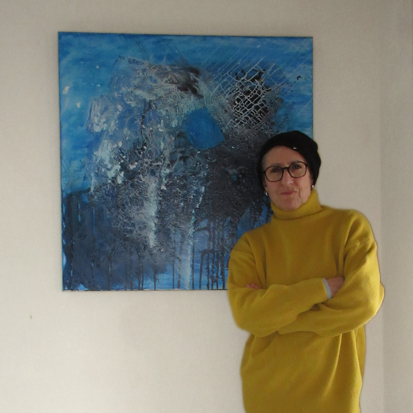 Person in gelbem Rollkragenpullover steht mit verschränkten Armen vor einem großformatigen abstrakten Gemälde in Blau- und Grautönen.