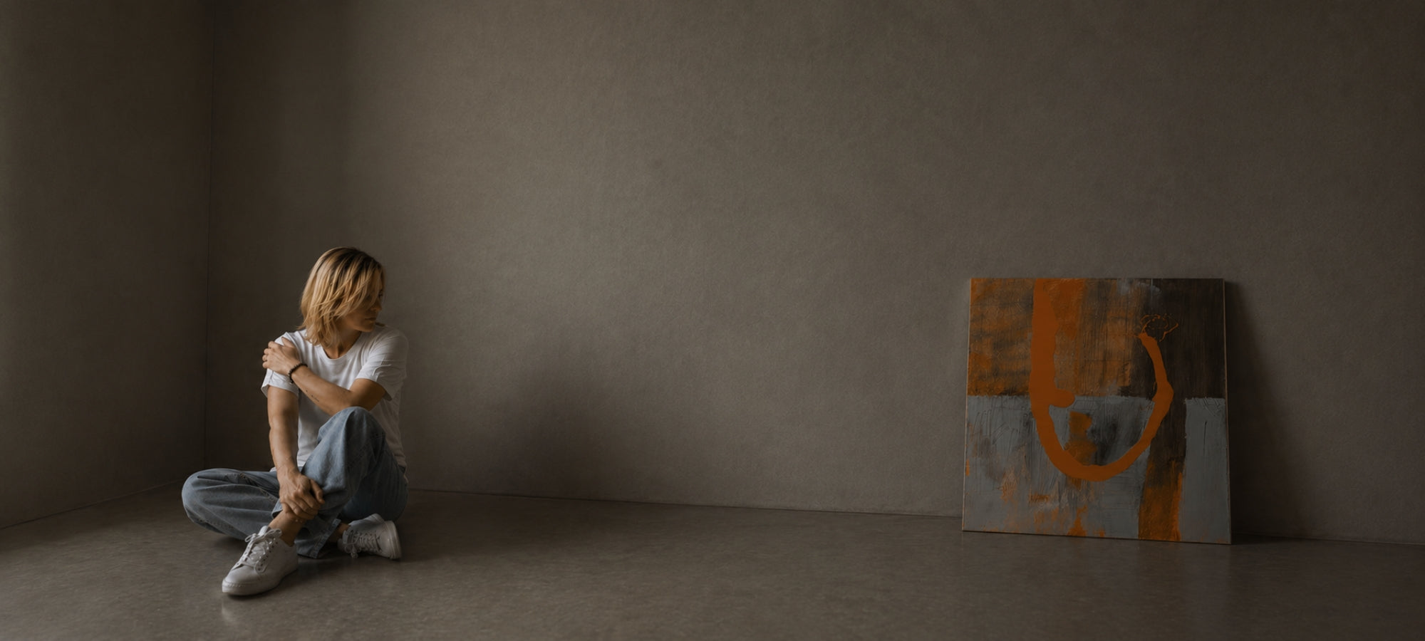 Eine Künstlerin mit blonden Haaren sitzt in entspannter Pose auf dem Boden in einer Ecke eines minimalistisch gestalteten, kargen Raumes mit grauen Wänden. Sie trägt ein weißes T-Shirt, Bluejeans und weiße Sneaker. Rechts von ihr lehnt ein quadratisches, abstraktes Gemälde an der Wand, das durch markante orangefarbene Pinselstriche auf grau-braunem Grund besticht. Die Szene ist ruhig und in gedämpftem, natürlichem Licht gehalten.