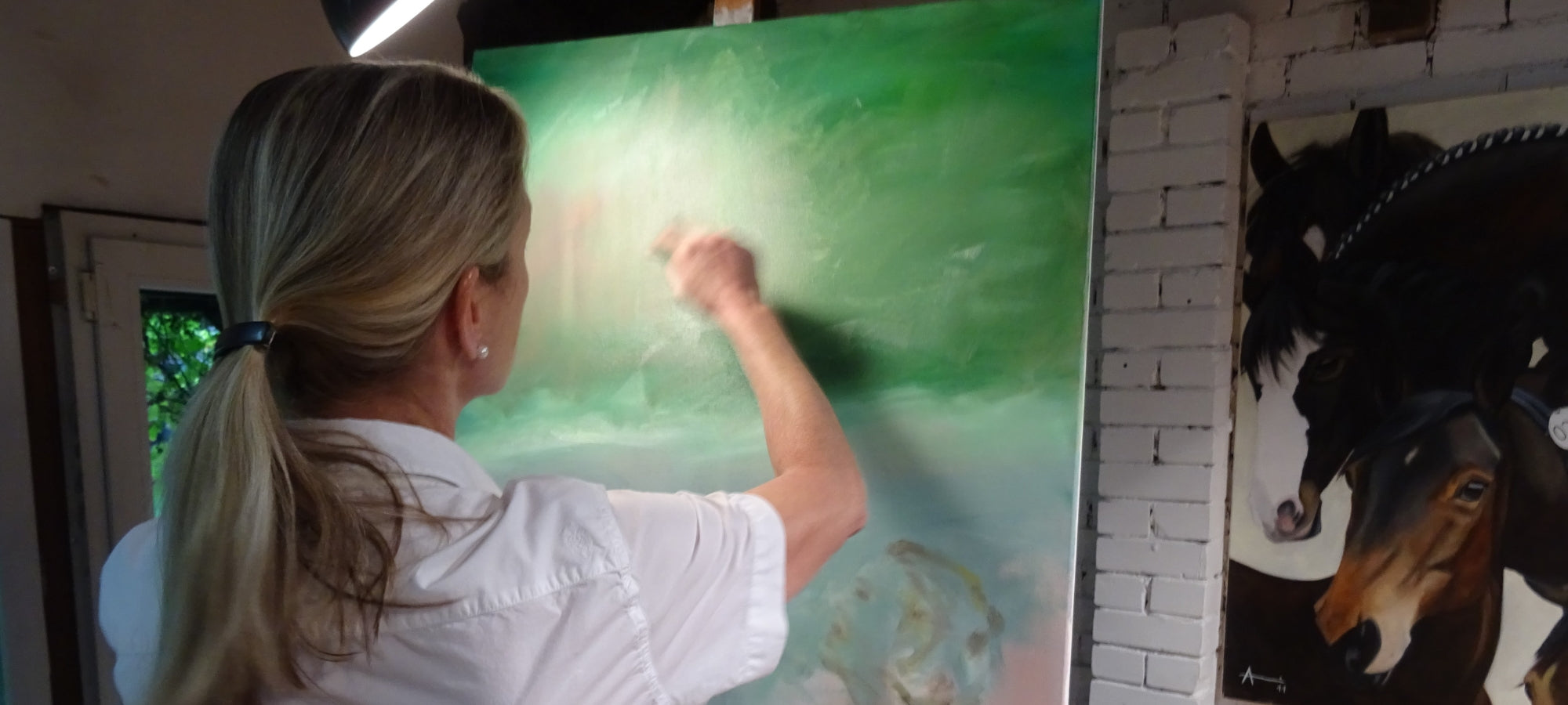 Eine blonde Frau von hinten, die vor einer grünen Leinwand steht und mit der rechten Hand malt. Links ist eine Tür zu sehen, rechts ein Teil eines anderen Gemäldes mit Pferdeköpfen. Es handelt sich um die Künstlerin Anke Dinkelbach von ARTSPACER.