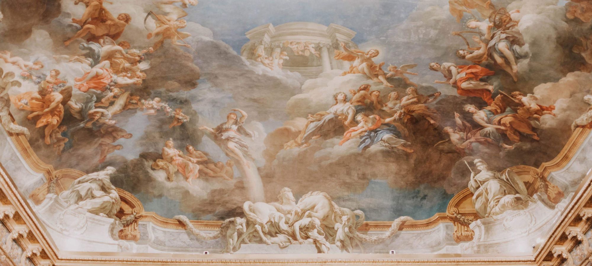 Klassisches barockes Deckenfresko mit mythologischen Figuren und Engeln in einem Wolkenhimmel, gehalten in sanften Pastelltönen mit goldfarbenen architektonischen Details.