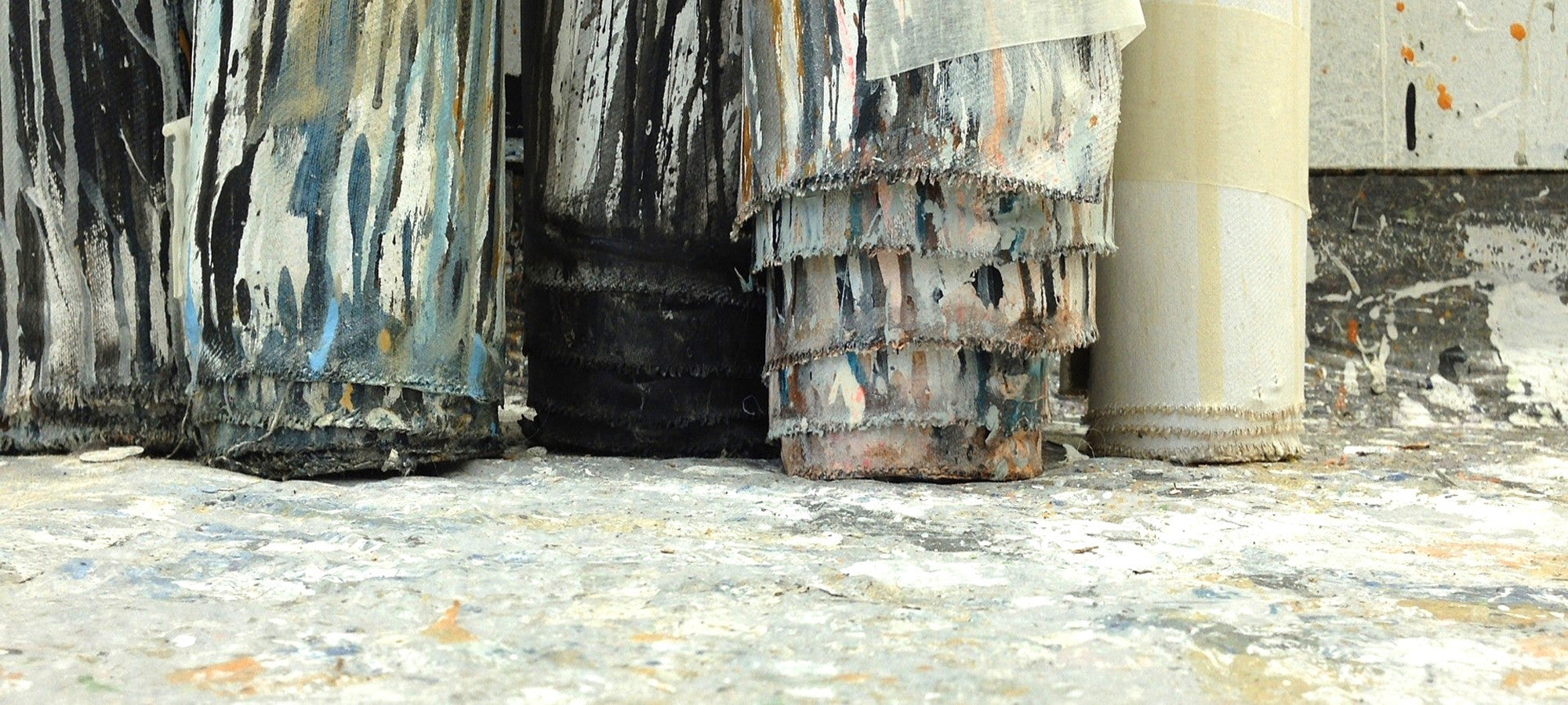 Mehrere großformatige, abstrakt bemalte Leinwände stehen auf einem farbverschmierten Atelierboden; dicke Farbschichten, vertikale Schwarz- und Blautöne sowie sichtbare Stoffkanten und aufgerollte Leinwand verleihen dem Werk eine rohe, materialbetonte Wirkung.