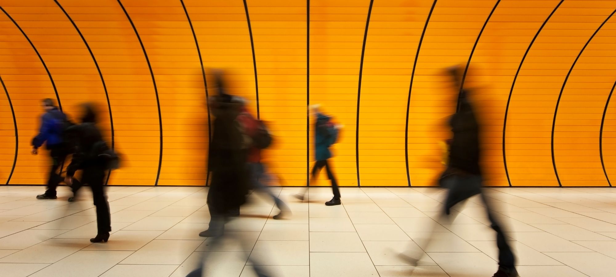 Street-Photography in einer U-Bahn-Station: Verschwommene Silhouetten von Menschen in Bewegung vor einer leuchtend gelben, grafisch strukturierten Wand mit schwarzen Linien.