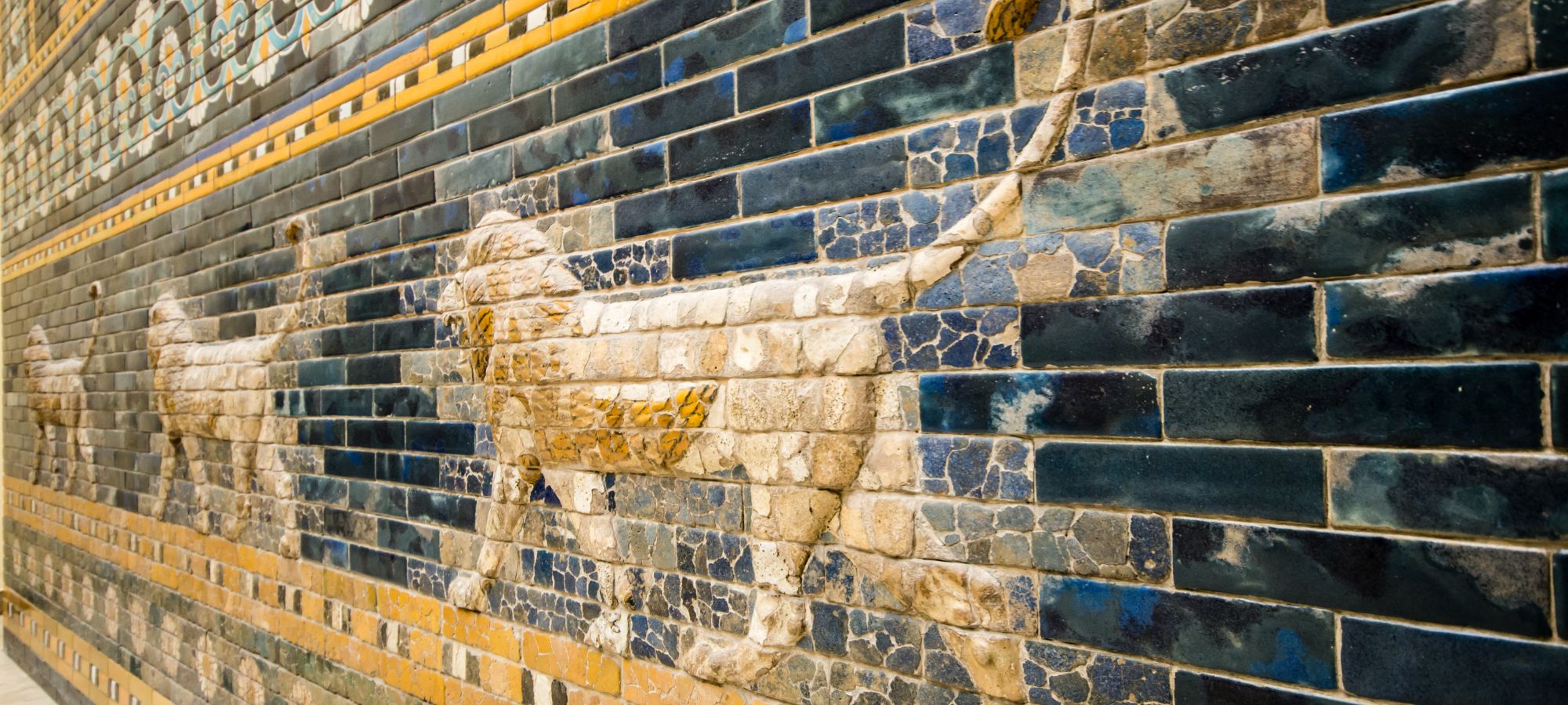 Antikes Wandmosaik aus glasierten Ziegeln mit schreitenden Löwen in Relief-Optik auf tiefblauem Hintergrund mit ockerfarbenen Ornamentbändern.