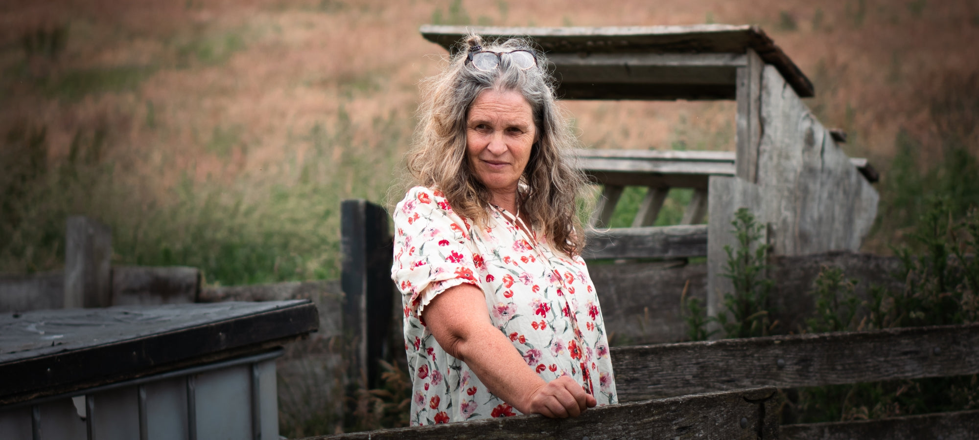 Person mit lockigem, schulterlangem Haar steht im Freien vor alten Holzkonstruktionen; trägt eine geblümte Bluse und lehnt an einem Zaunelement in ländlicher Umgebung.
