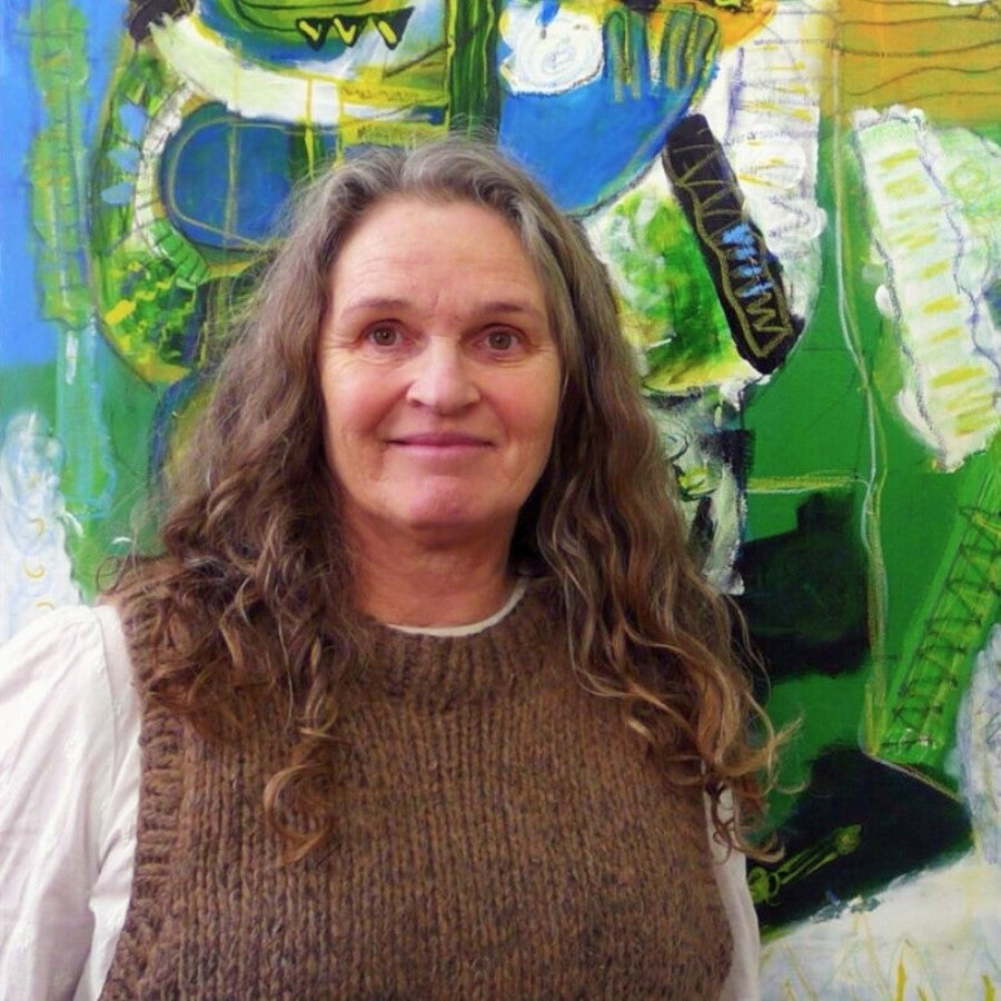 Person mit langen, gewellten Haaren steht vor einem großformatigen, farbintensiven abstrakten Gemälde in Grün‑, Blau‑ und Gelbtönen; trägt einen braunen Strickpullover über einem hellen Oberteil.