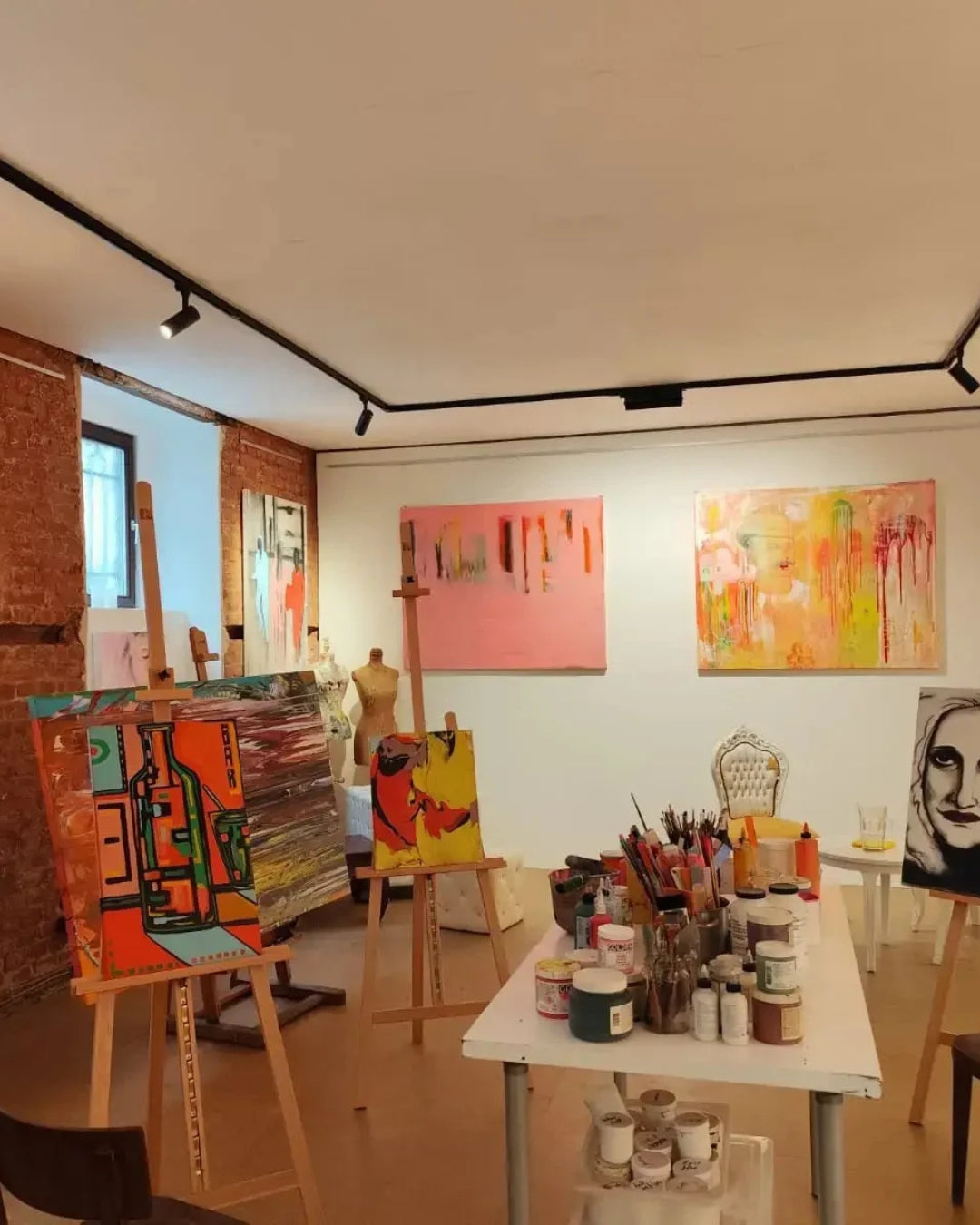Künstleratelier mit mehreren abstrakten und figurativen Gemälden auf Staffeleien und an den Wänden, Arbeitstisch mit Farben und Pinseln sowie freigelegter Backsteinwand.
