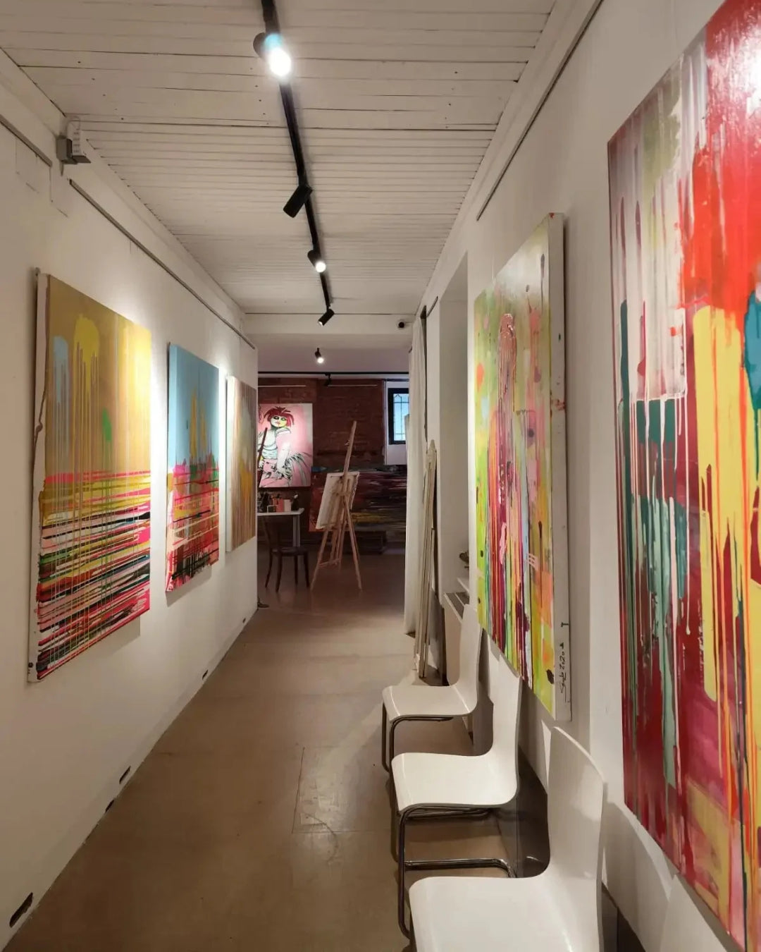Heller Galeriegang mit mehreren großformatigen, farbintensiven Gemälden an weißen Wänden, schwarzen Deckenspots, drei weißen Stühlen und einem Atelierbereich mit Staffelei im Hintergrund.