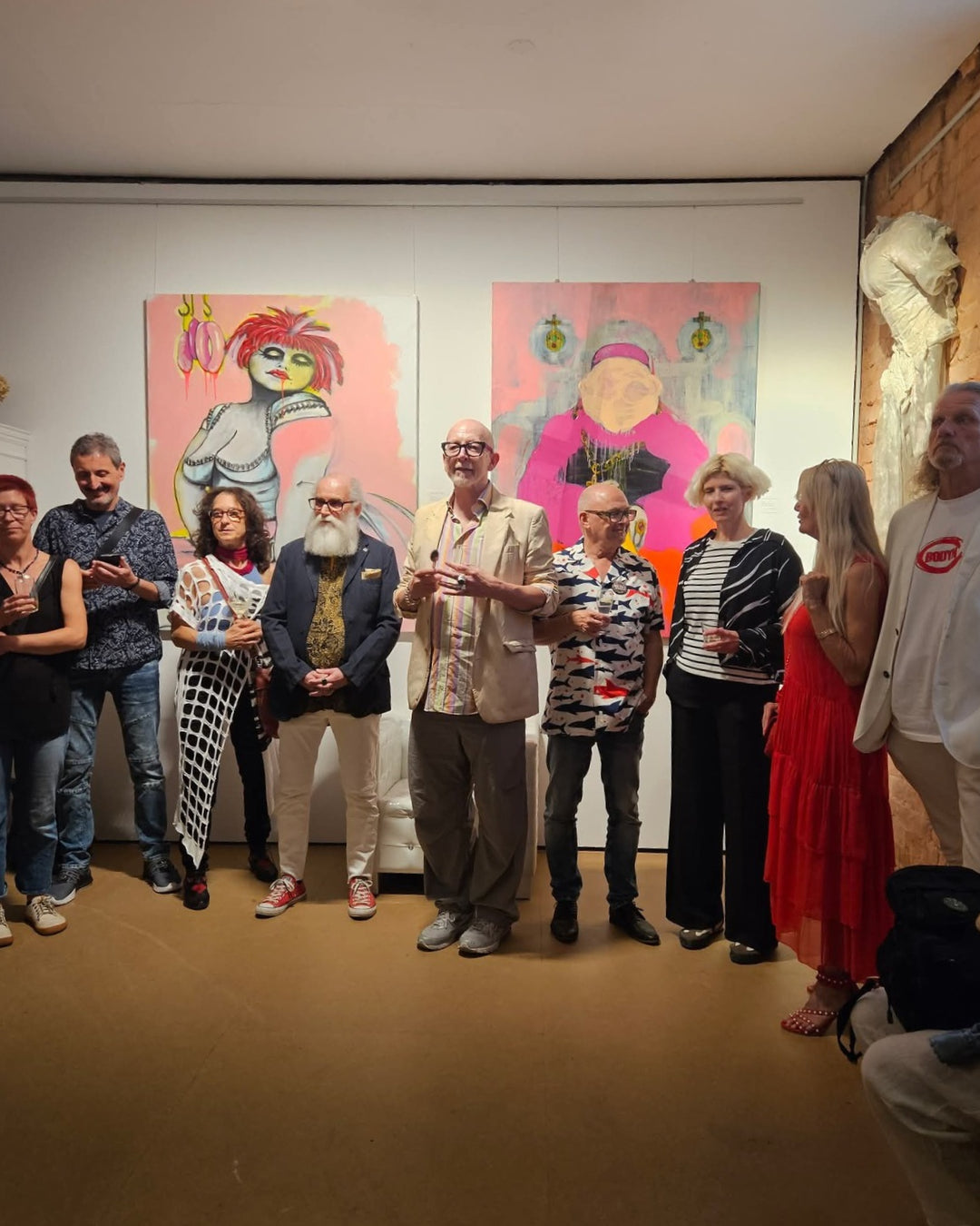 Gruppenaufnahme bei einer Kunstausstellung vor zwei großformatigen figurativen Gemälden in Rosa, Pink, Grau und Schwarz sowie einer weißen Skulptur an der Backsteinwand.