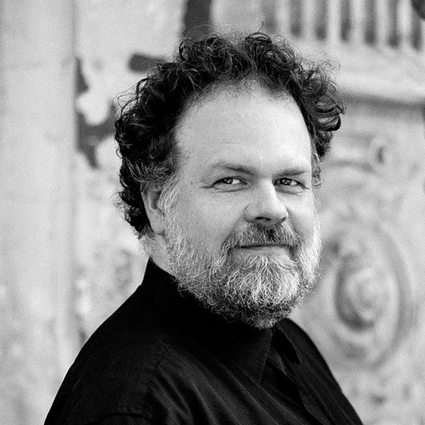 Schwarz-Weiß-Porträt des Künstlers Jens Joneleit mit lockigem Haar und Bart, Seitenprofil vor dezentem architektonischem Hintergrund.