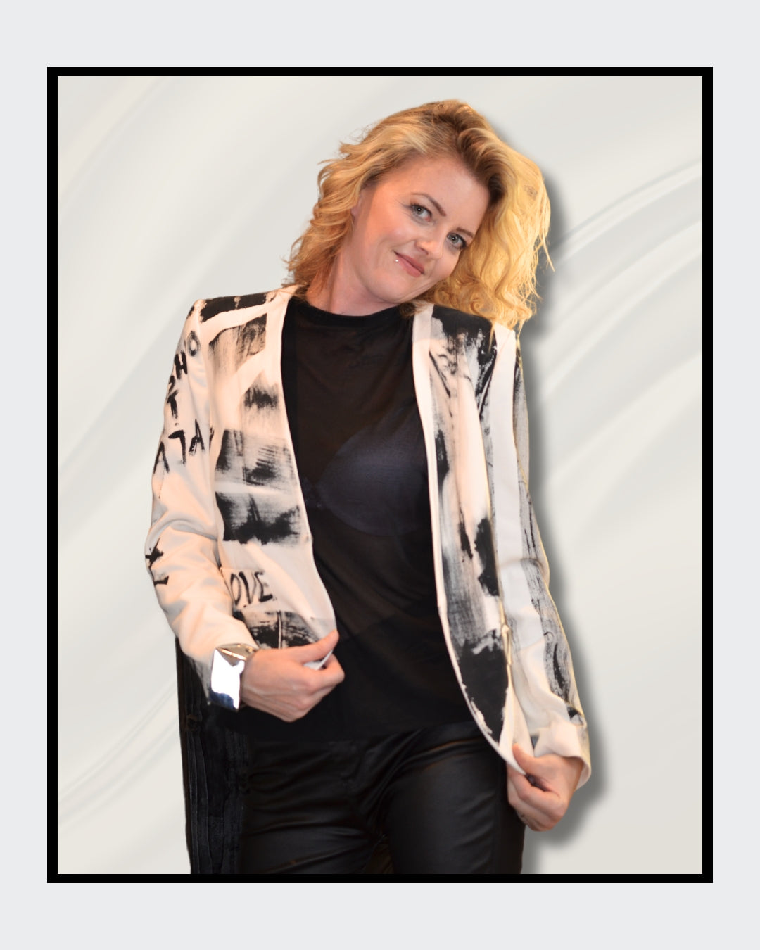 Eine blonde Frau steht vor einem cremefarbenen Hintergrund und blickt nach rechts. Sie trägt einen weißen Blazer mit schwarzen, abstrakten Mustern und Schriftzügen, darunter das Wort "LOVE". Darunter trägt sie ein schwarzes Oberteil und eine schwarze Hose. Fashionart von Kaja Schäfer, exklusiv bei ARTSPACER, Online-Galerie.
