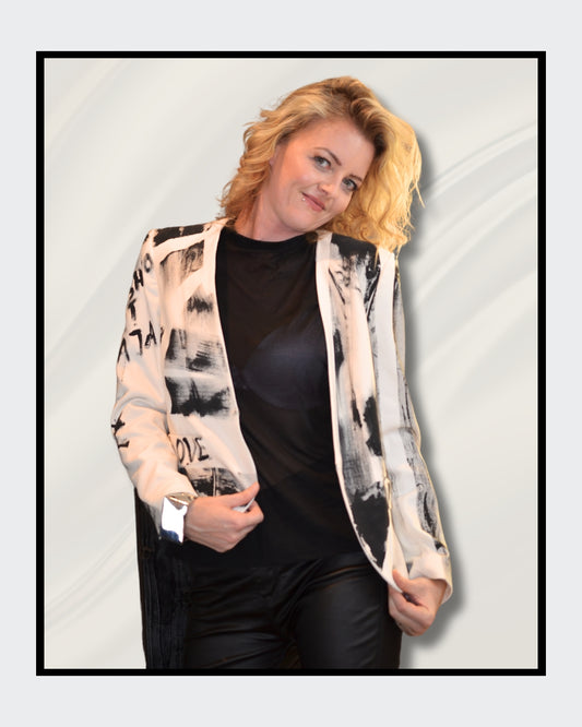 Eine blonde Frau steht vor einem cremefarbenen Hintergrund und blickt nach rechts. Sie trägt einen weißen Blazer mit schwarzen, abstrakten Mustern und Schriftzügen, darunter das Wort "LOVE". Darunter trägt sie ein schwarzes Oberteil und eine schwarze Hose. Fashionart von Kaja Schäfer, exklusiv bei ARTSPACER, Online-Galerie.
