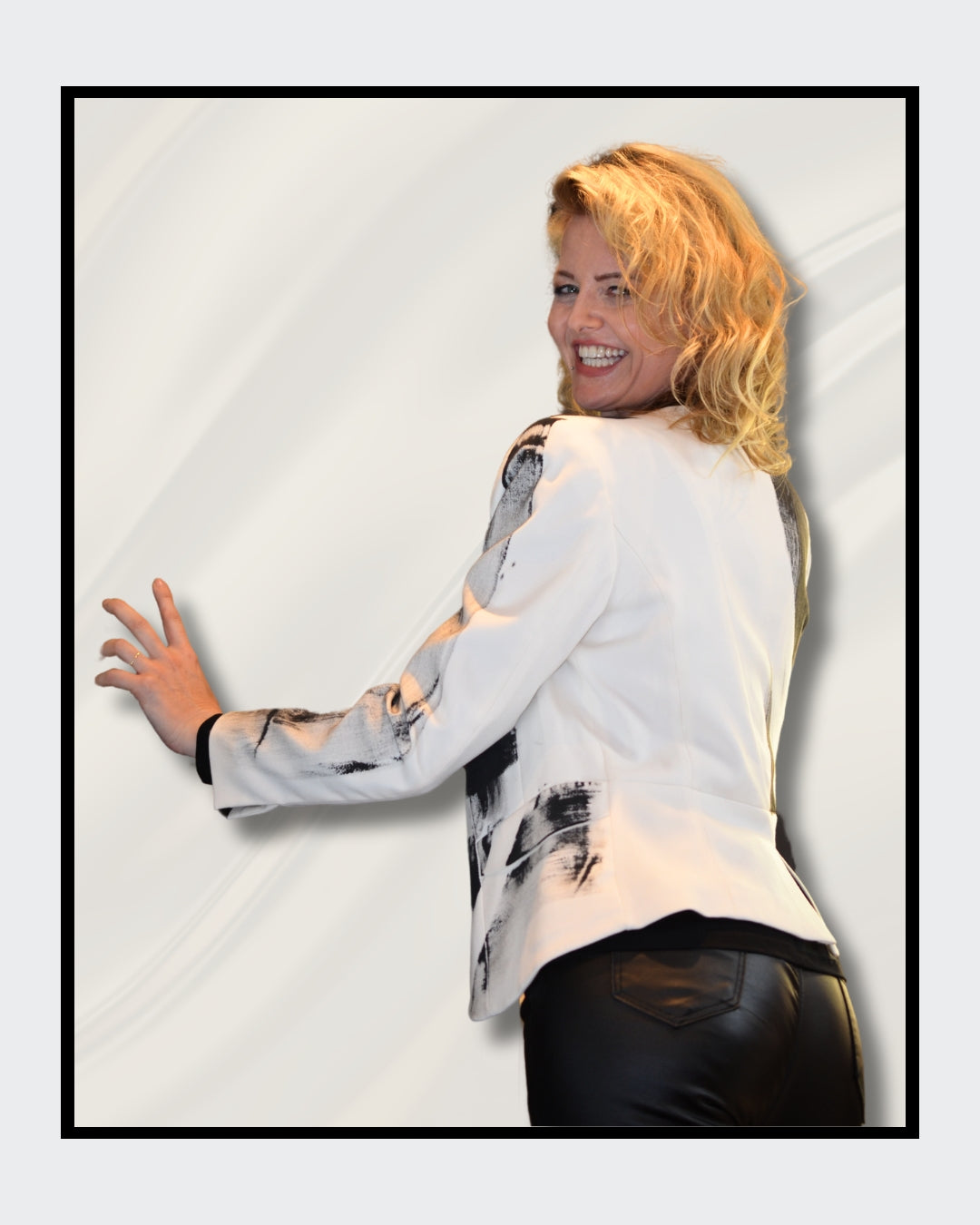 Eine blonde Frau steht vor einem cremefarbenen Hintergrund und blickt nach rechts. Sie trägt einen weißen Blazer mit schwarzen, abstrakten Mustern und Schriftzügen, darunter das Wort "LOVE". Darunter trägt sie ein schwarzes Oberteil und eine schwarze Hose. Fashionart von Kaja Schäfer, exklusiv bei ARTSPACER, Online-Galerie.
