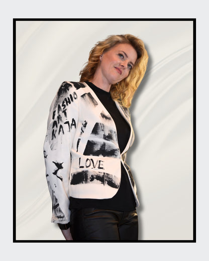 Eine blonde Frau steht vor einem cremefarbenen Hintergrund und blickt nach rechts. Sie trägt einen weißen Blazer mit schwarzen, abstrakten Mustern und Schriftzügen, darunter das Wort "LOVE". Darunter trägt sie ein schwarzes Oberteil und eine schwarze Hose. Fashionart von Kaja Schäfer, exklusiv bei ARTSPACER, Online-Galerie.
