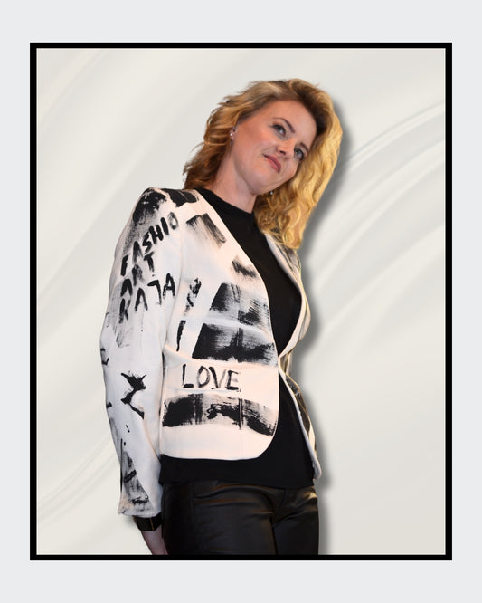 Eine blonde Frau steht vor einem cremefarbenen Hintergrund und blickt nach rechts. Sie trägt einen weißen Blazer mit schwarzen, abstrakten Mustern und Schriftzügen, darunter das Wort "LOVE". Darunter trägt sie ein schwarzes Oberteil und eine schwarze Hose. Fashionart von Kaja Schäfer, exklusiv bei ARTSPACER, Online-Galerie.
