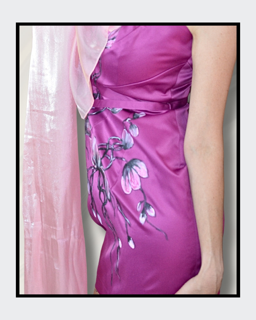 Ein elegantes, taillenbetontes Damenkleid in einem satten Purpurton. Der Stoff ist aus glänzendem Satin und mit zarten, handgemalten Blüten in verschiedenen Rosa- und Weißtönen verziert. Die Blütenranken schlängeln sich über das Kleid und verleihen ihm einen romantischen Touch. Ein zartes, durchscheinendes Tuch in einem helleren Rosaton liegt über einer Schulter und ergänzt den femininen Look.  Handbemalt von Kaja Schäfer.