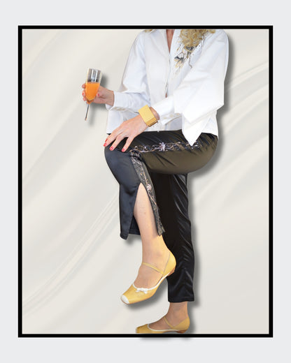 Fashionart von Kaja Schäfer für ARTSPACER: Die untere Hälfte einer Frau in einer eleganten Pose. Schwarze Satinhose mit silbernen und grauen Linien, gelbe Ballerinas und ein orangefarbenes Getränk in der Hand. Cremiger Hintergrund mit fließenden Stoffen.