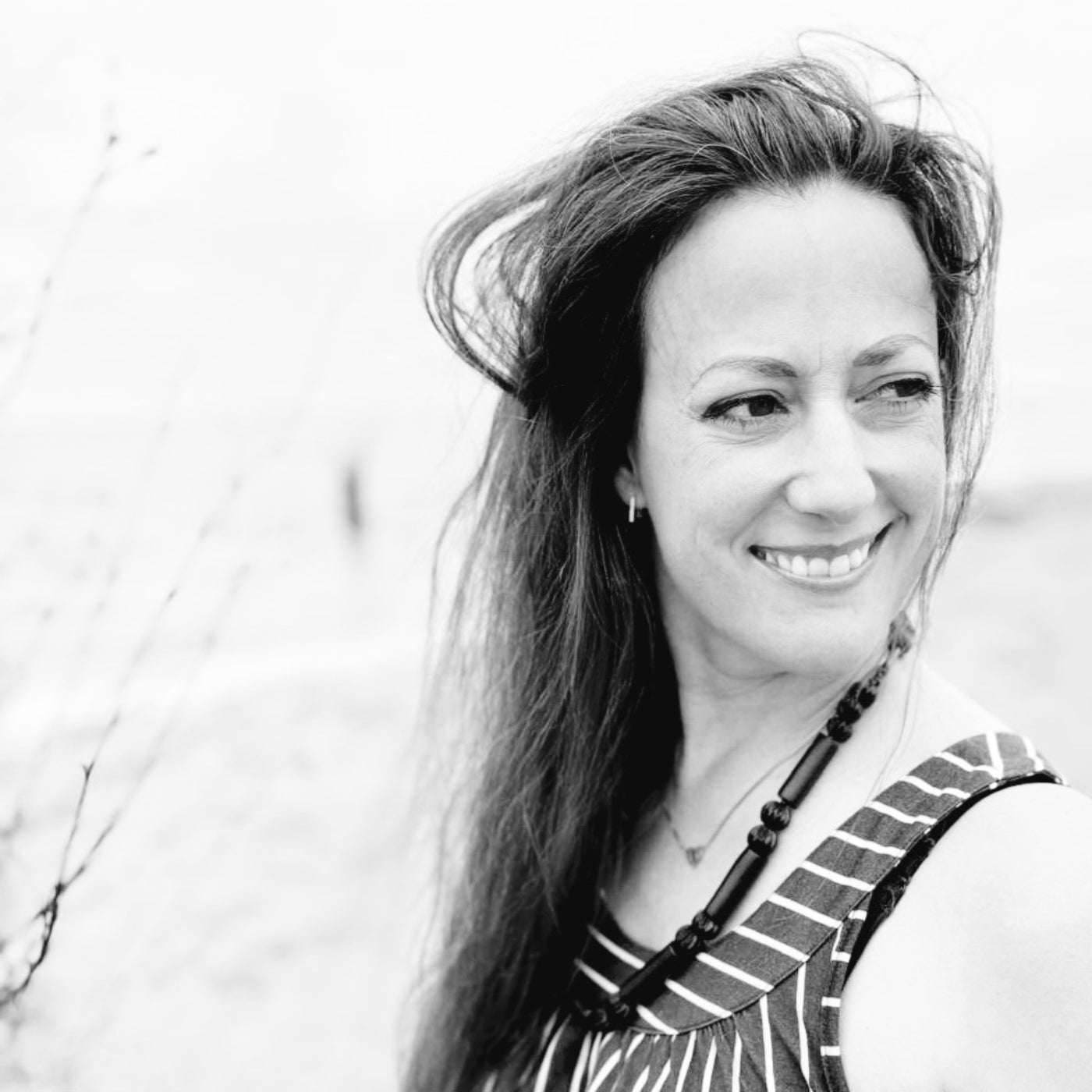 Ein Schwarz-Weiß-Porträt der Künstlerin Melanie Zürn im Freien. Sie blickt lächelnd zur Seite, wobei ihr langes, dunkles Haar leicht vom Wind verweht wird. Sie trägt ein gestreiftes Oberteil und eine markante, dunkle Halskette aus großen Gliedern. Der Hintergrund ist hell und verschwommen, was eine natürliche und lebendige Atmosphäre schafft.