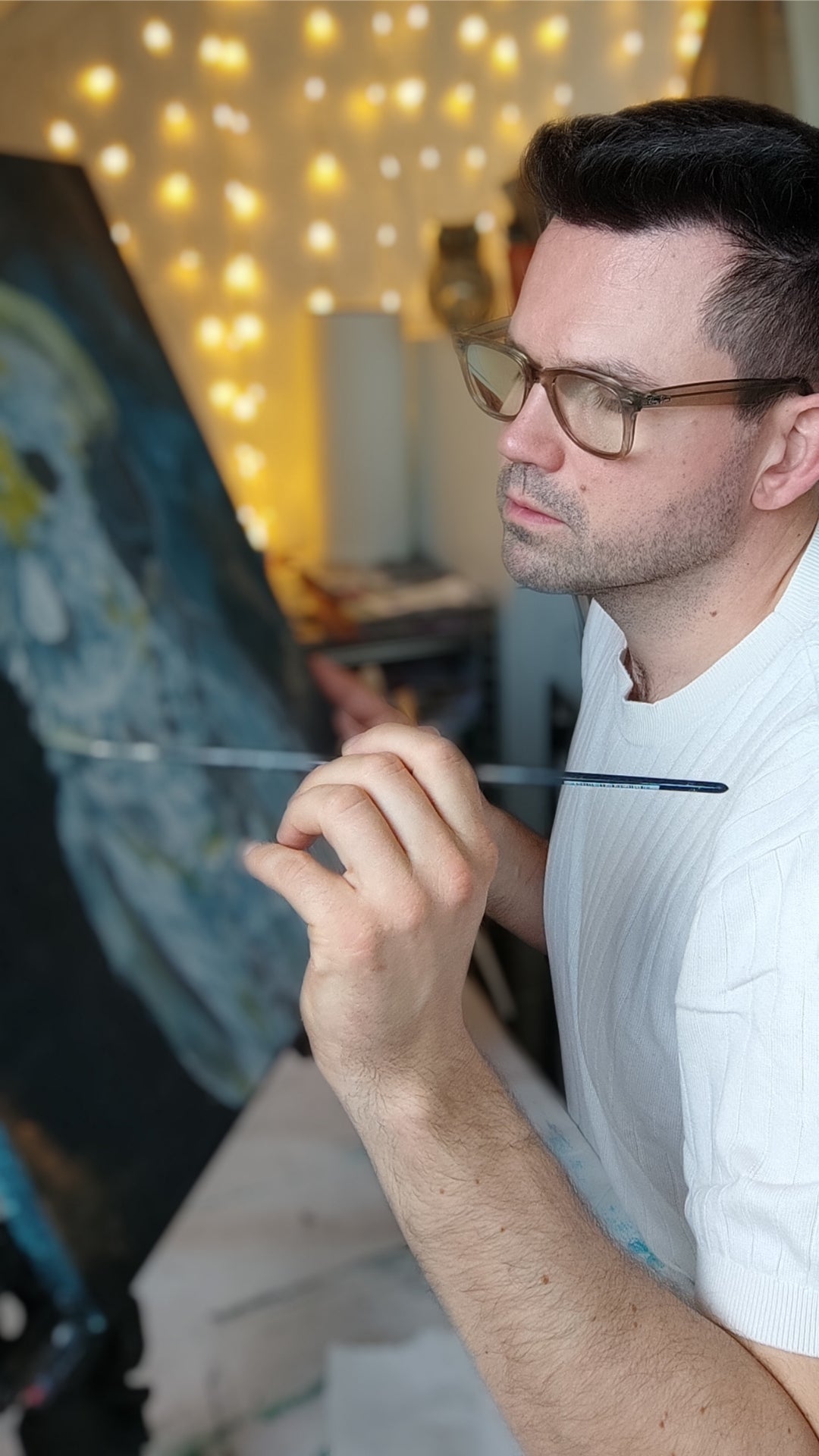 Nahaufnahme eines Künstlers im Profil, der konzentriert mit einem feinen Pinsel an einer dunklen Leinwand arbeitet. Im Hintergrund sind warme Lichterketten zu sehen.