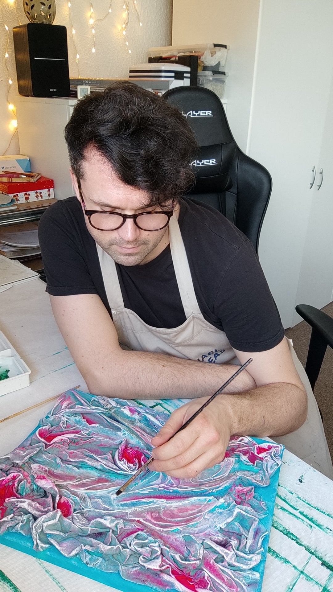 Der Künstler sitzt an einem Tisch und arbeitet mit einem Pinsel an einem strukturierten, abstrakten Kunstwerk in den Farben Pink, Blau und Weiß.