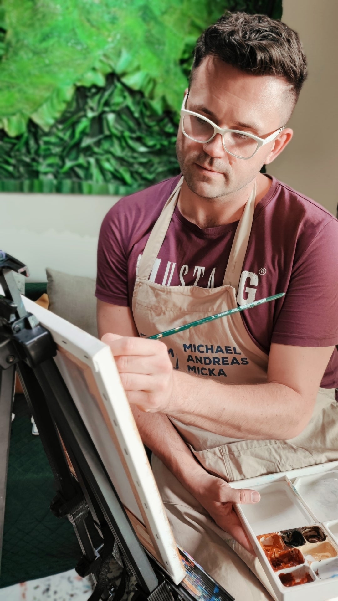 Michael Andreas Micka sitzt an einer Staffelei und malt konzentriert auf eine Leinwand. Er hält eine Farbpalette in der Hand und trägt seine Künstlerschürze.